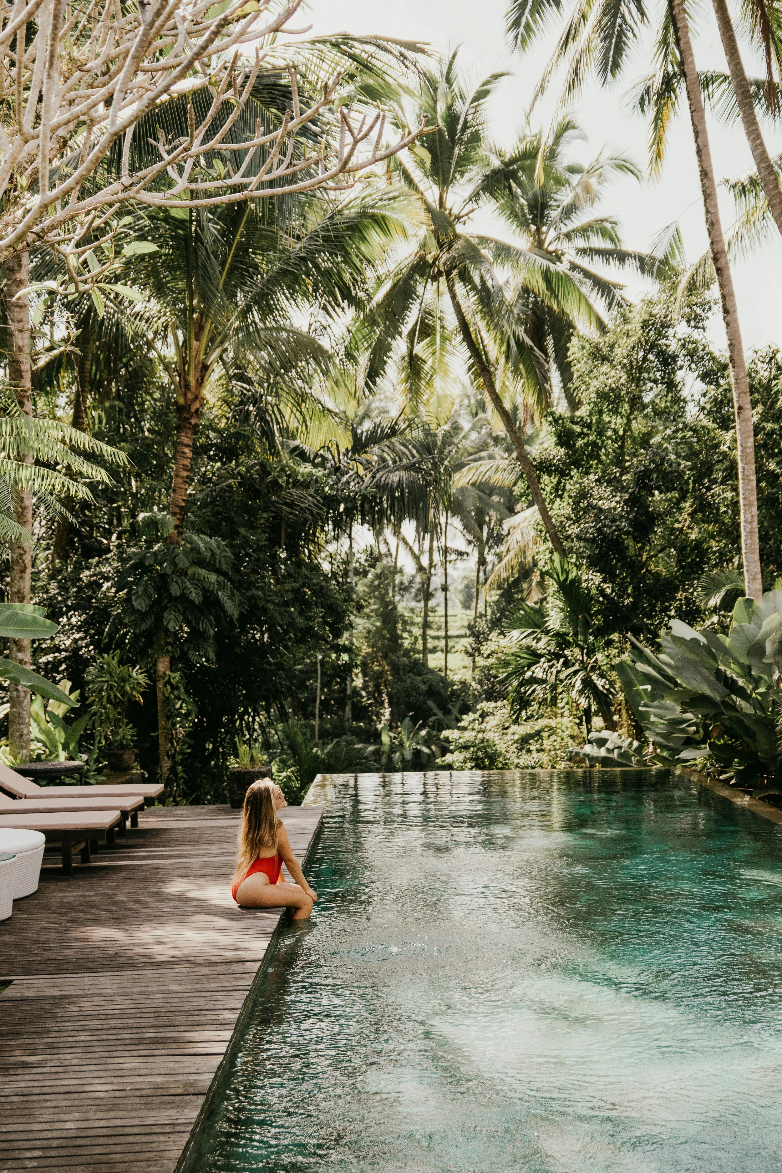 A woman in a red swimsuit sitting at the edge of an infinity pool surrounded by lush tropical greenery and tall palm trees.