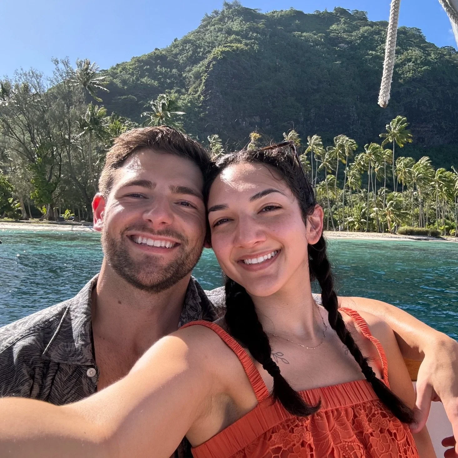 A smiling couple taking a selfie in front of a tropical beach with clear water, palm trees, and green mountains in the background.