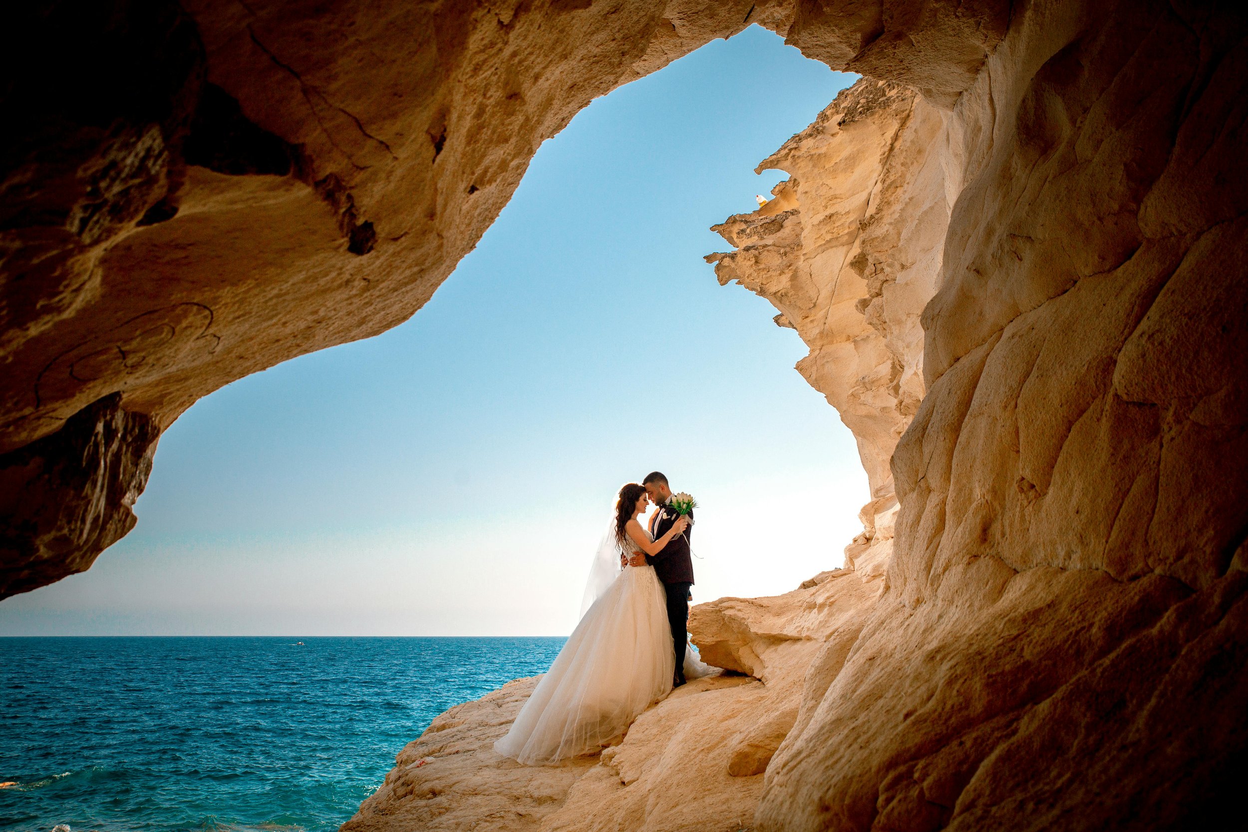 A bride and groom standing close together inside a natural rock formation, overlooking the ocean at sunset. The bride is in a white wedding dress, and the groom in a suit, holding a bouquet of flowers.