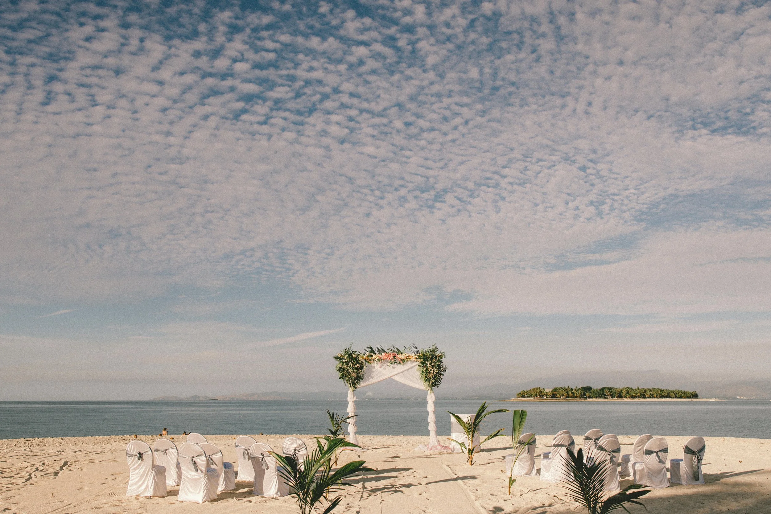 Beach wedding setup with chairs covered in white fabric, facing an arch decorated with flowers and greenery, seaside view with sand, ocean, small island, and partly cloudy sky.