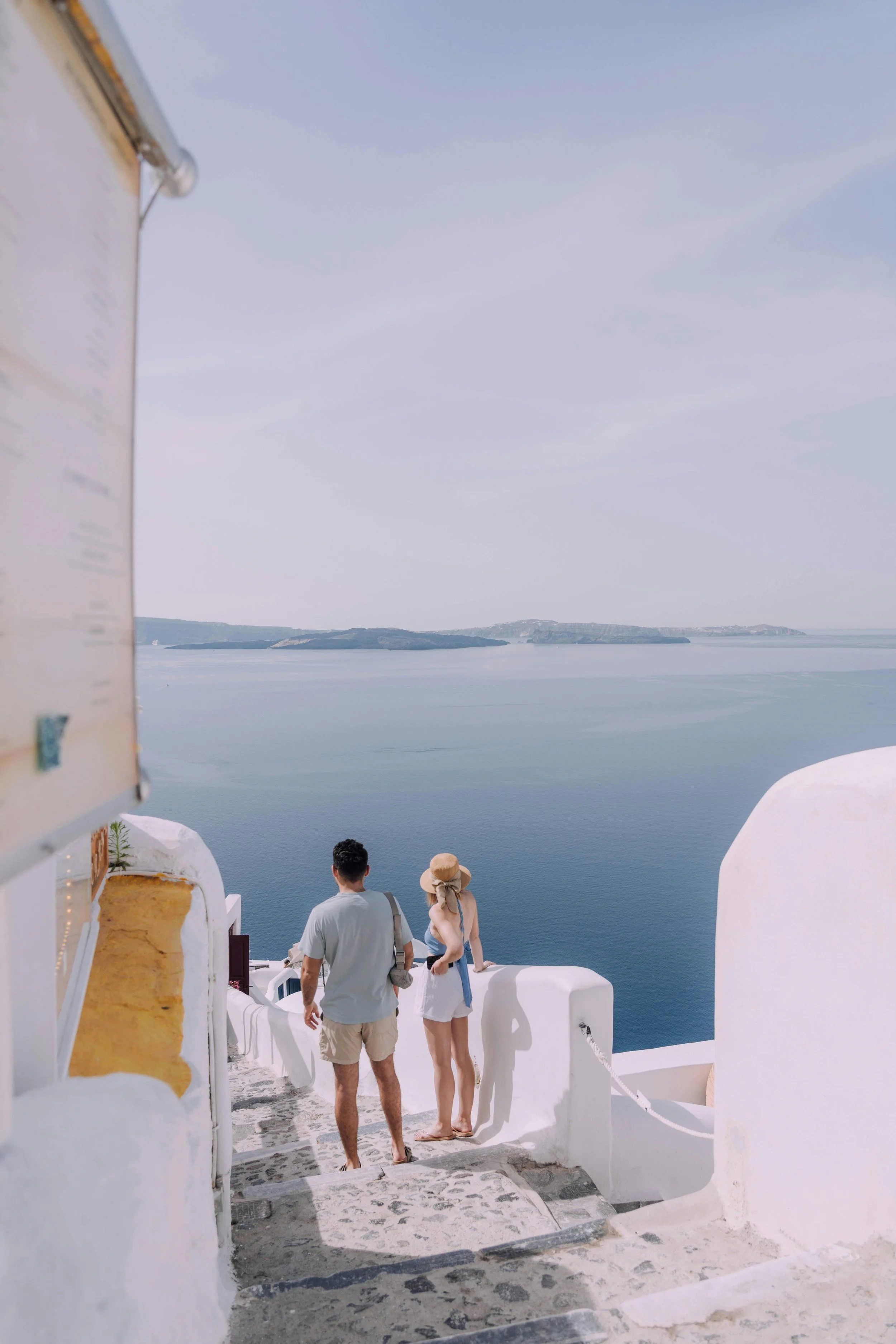Couple walking down white stairs in a Mediterranean coastal town, overlooking blue sea and distant islands, with whitewashed buildings and a clear sky.
