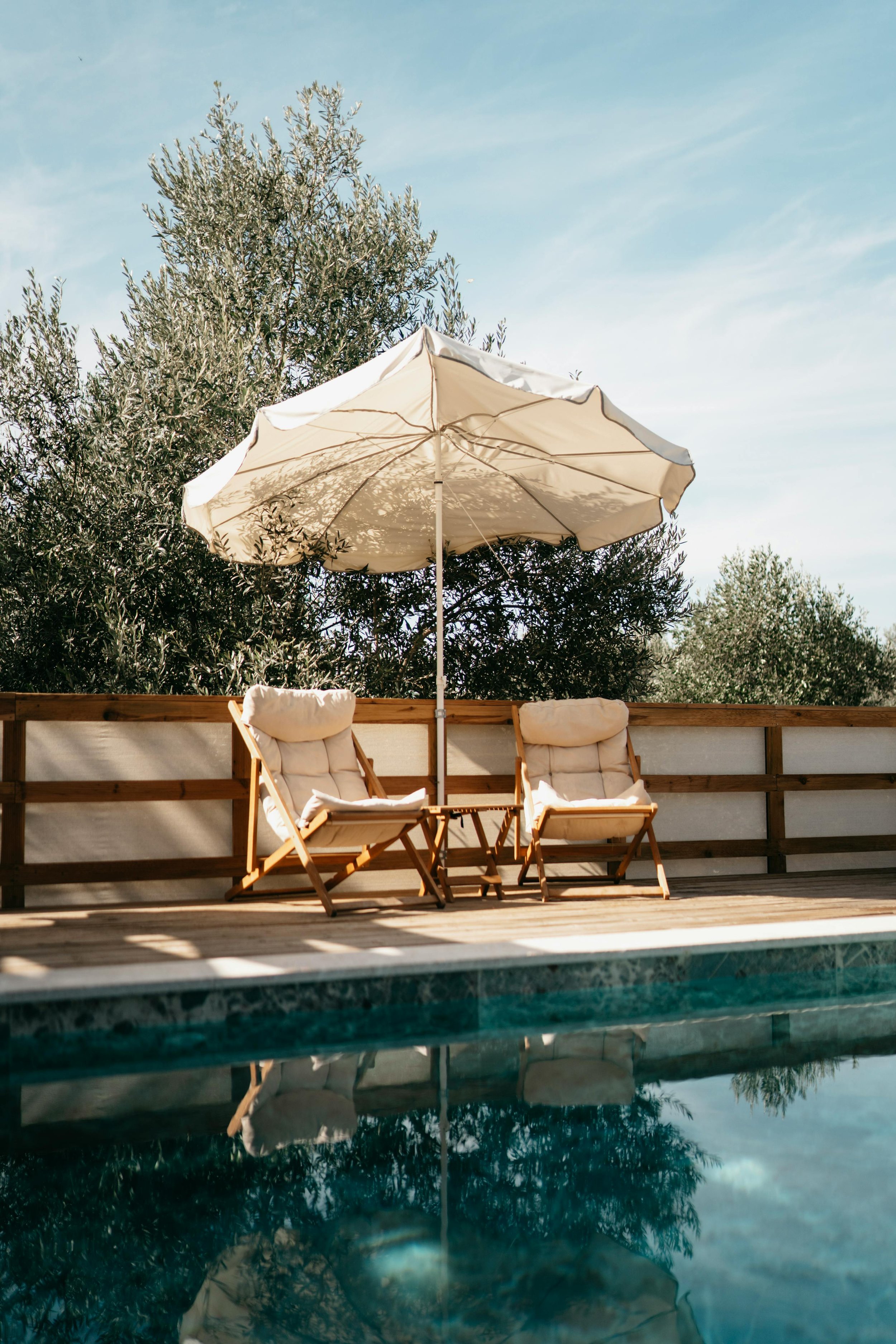 Poolside scene with two wooden lounge chairs with white cushions, a small matching table, a large white patio umbrella, a wooden fence, green trees, and a blue sky reflected in the swimming pool.