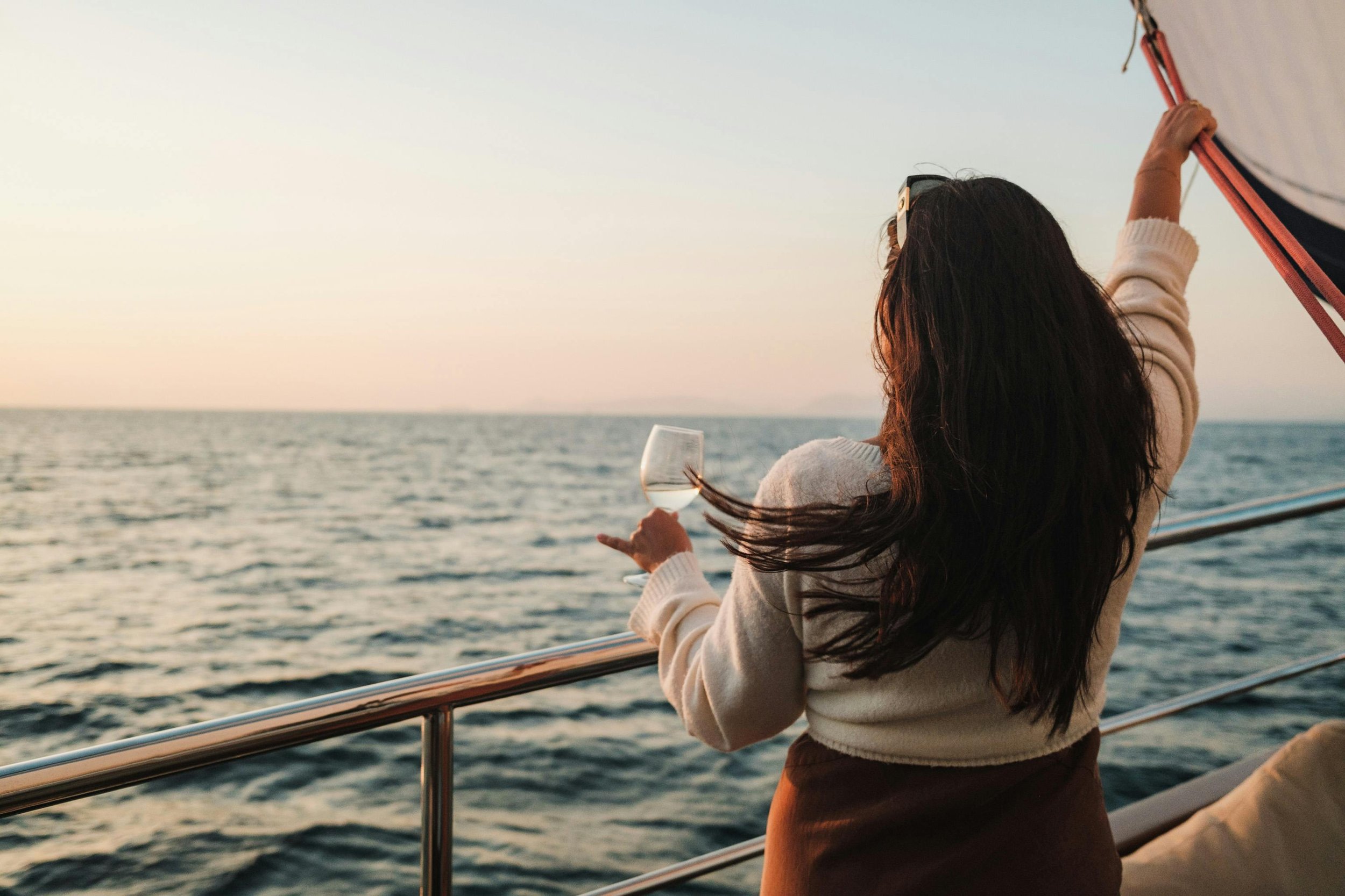 A woman with long dark hair holding a glass of white wine, standing on a boat during sunset, enjoying the ocean view.
