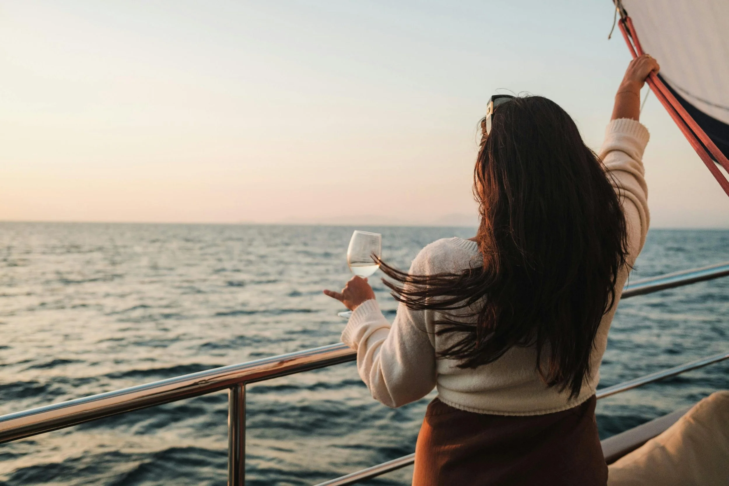 A woman with long dark hair holding a glass of white wine, standing on a boat during sunset, enjoying the ocean view.