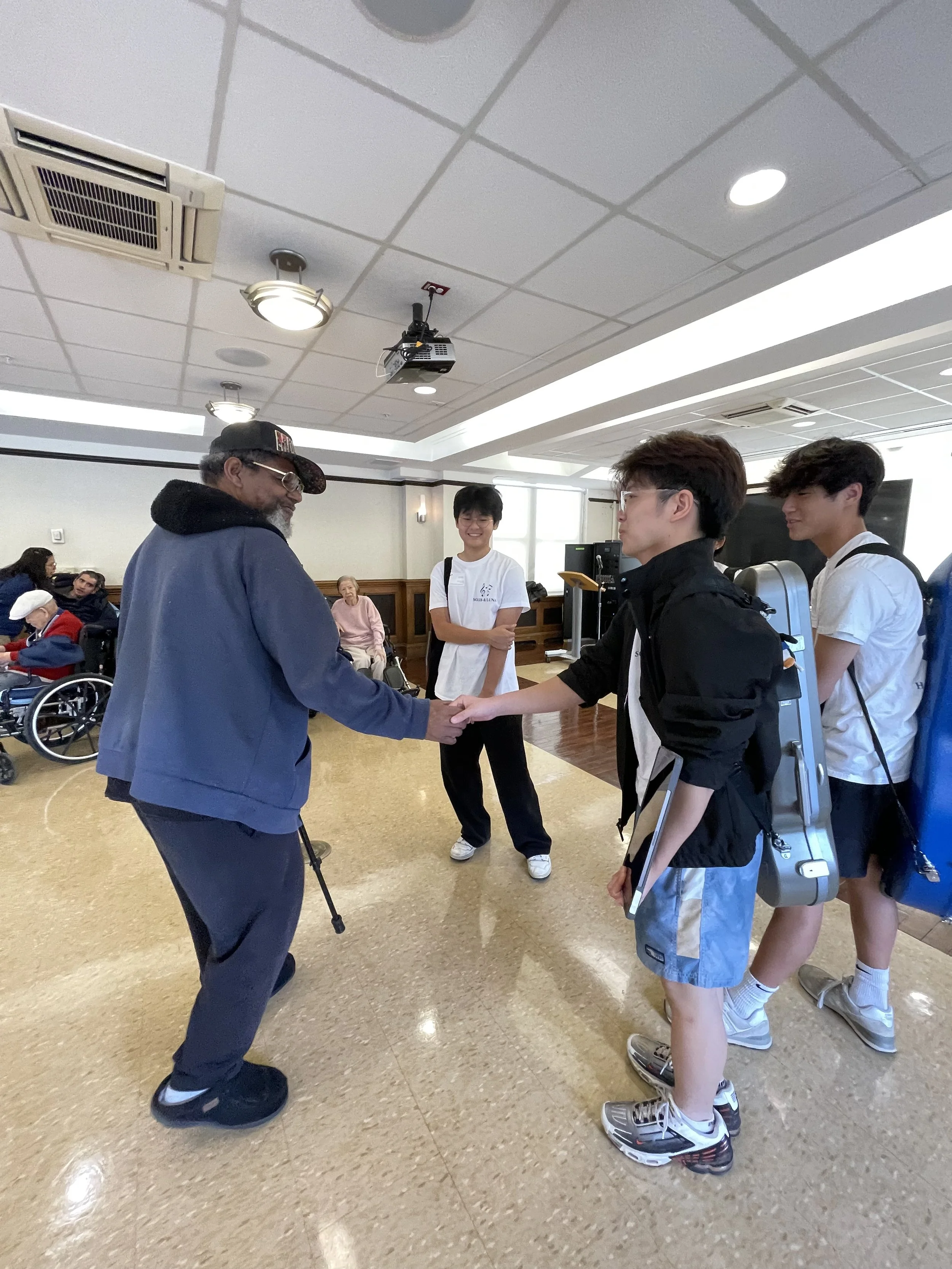 An indoor scene where a man is shaking hands with a young man carrying a guitar case, surrounded by three other young men, with elderly women seated in the background.