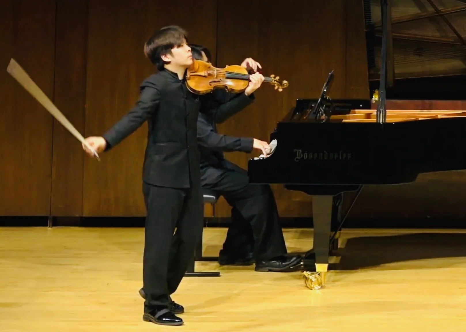 A young boy playing a violin while a woman plays a grand piano in a wooden-paneled room.