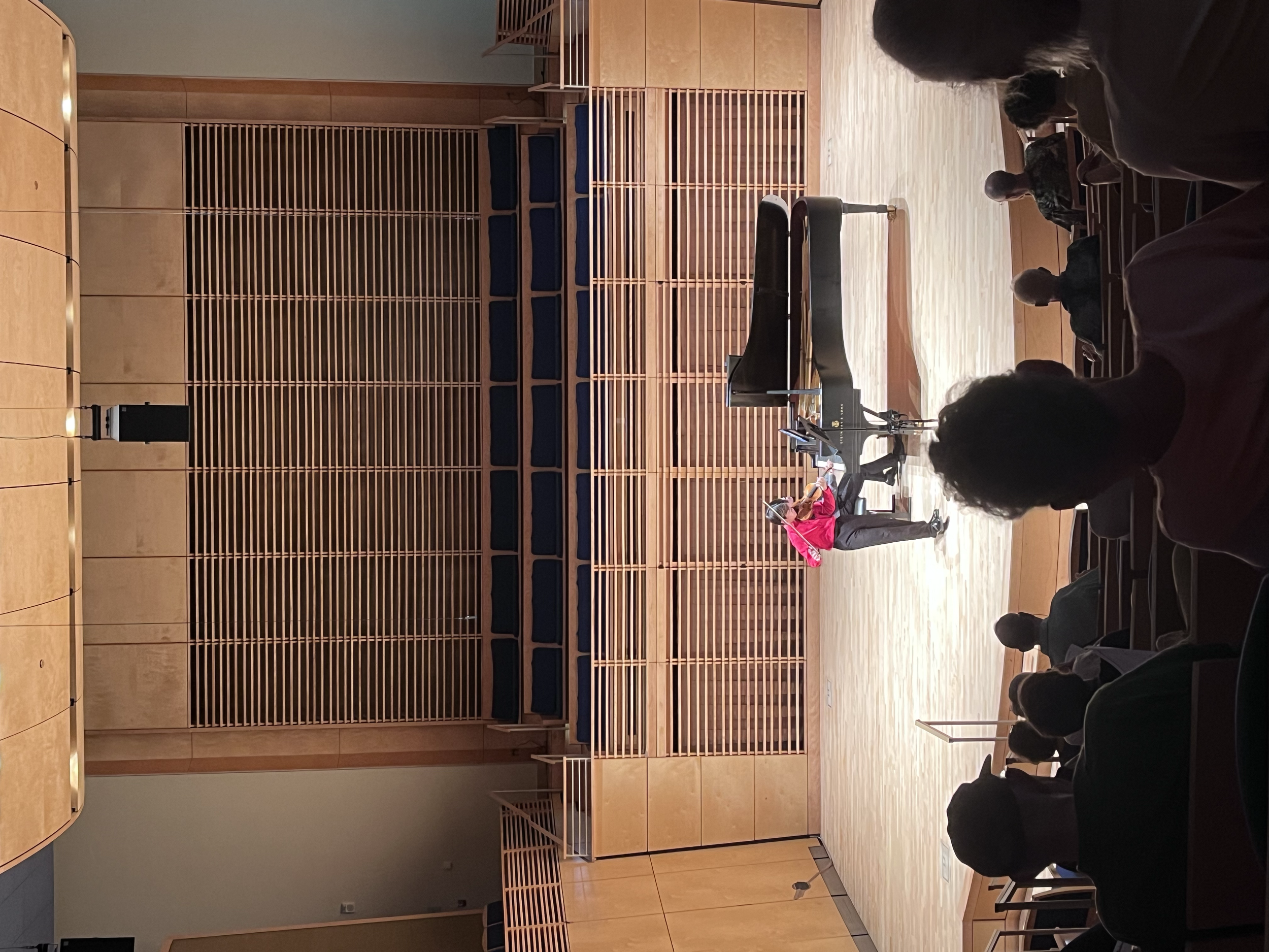 A woman in a red blazer performing on a grand piano on a stage in front of an audience in a concert hall with wood paneling and tiered seating.