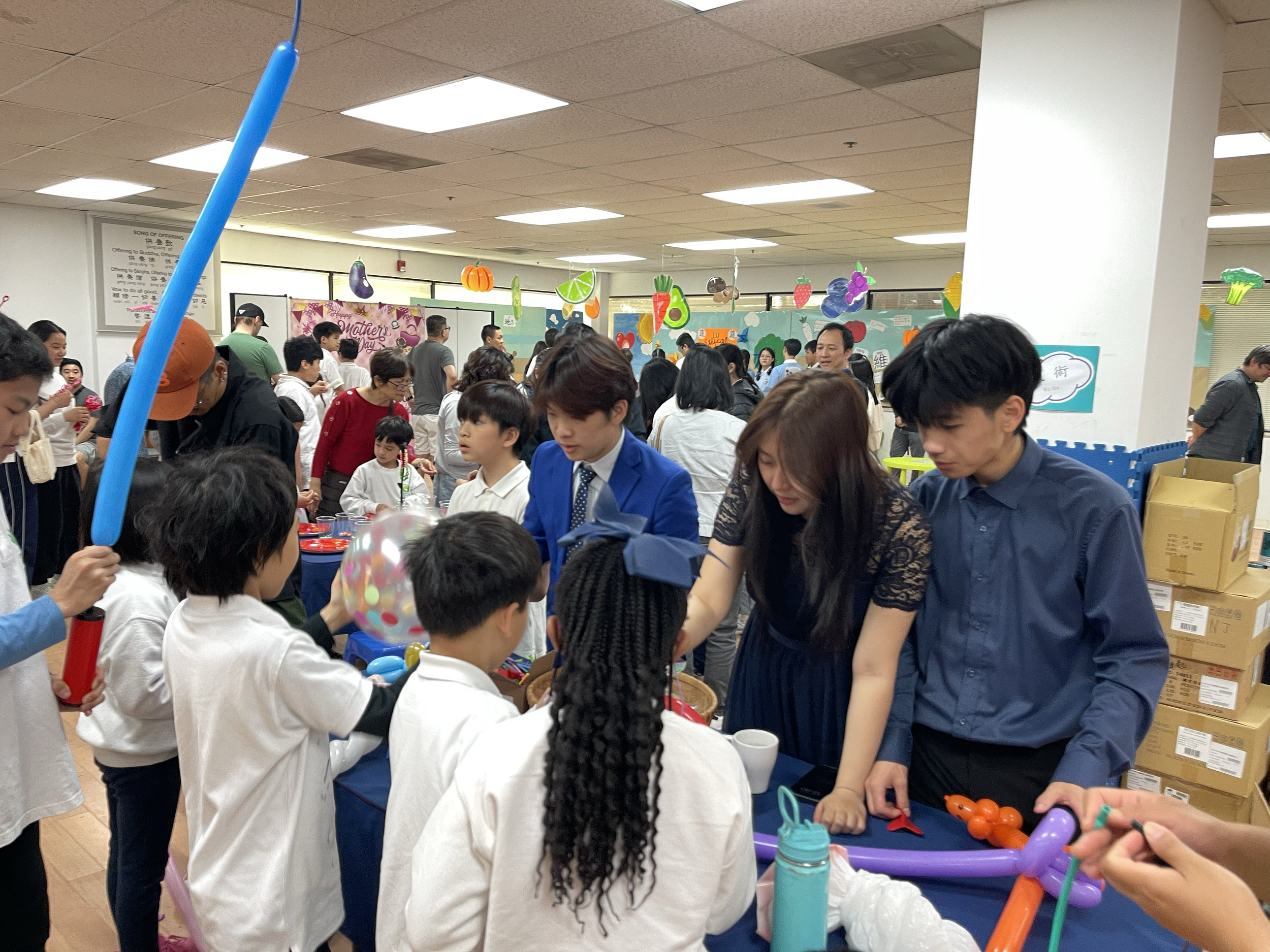 A crowded indoor event with children and adults, with decorations hanging from the ceiling, including pumpkins and fruit-shaped designs, and activities involving balloon twisting.