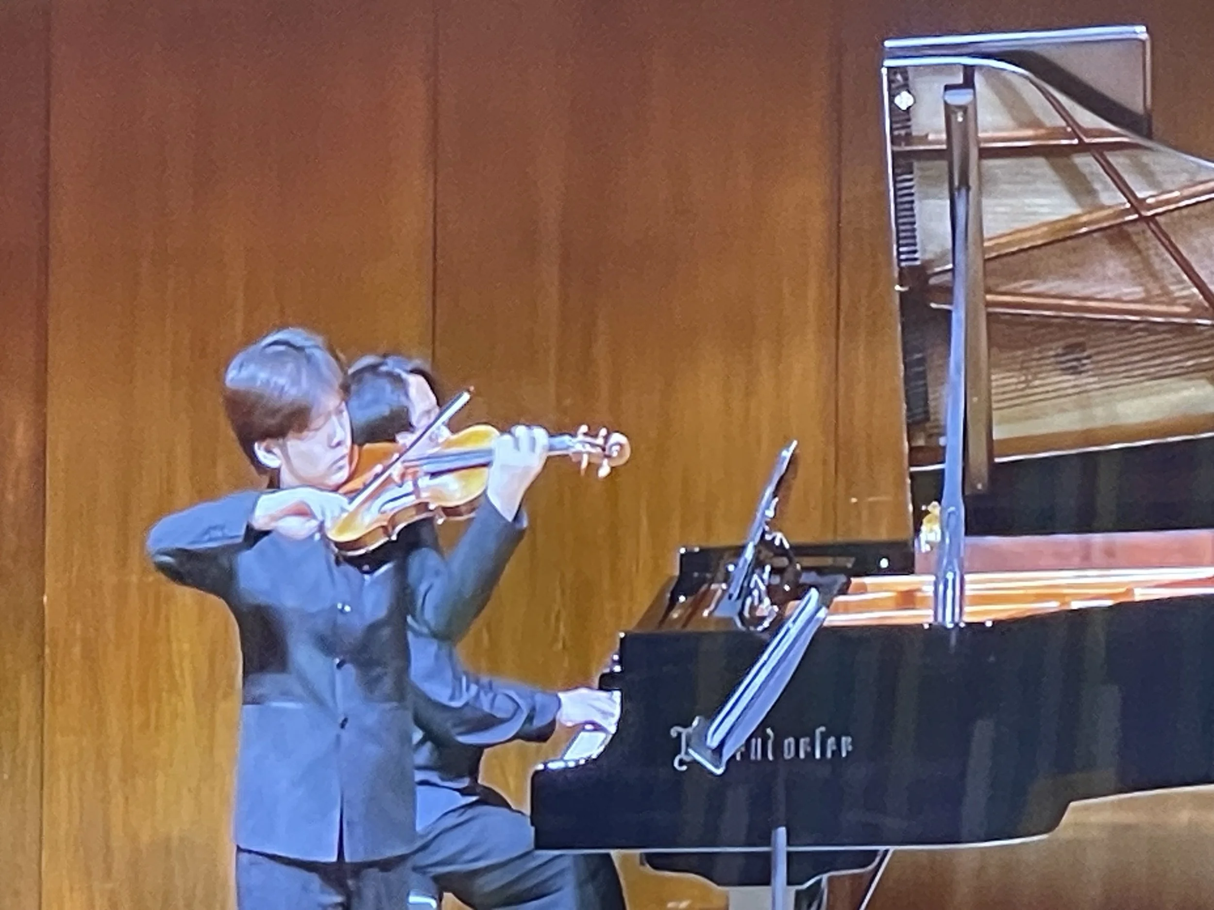 Two young musicians, a boy playing the violin and a girl playing the piano, perform on stage against a wooden background.