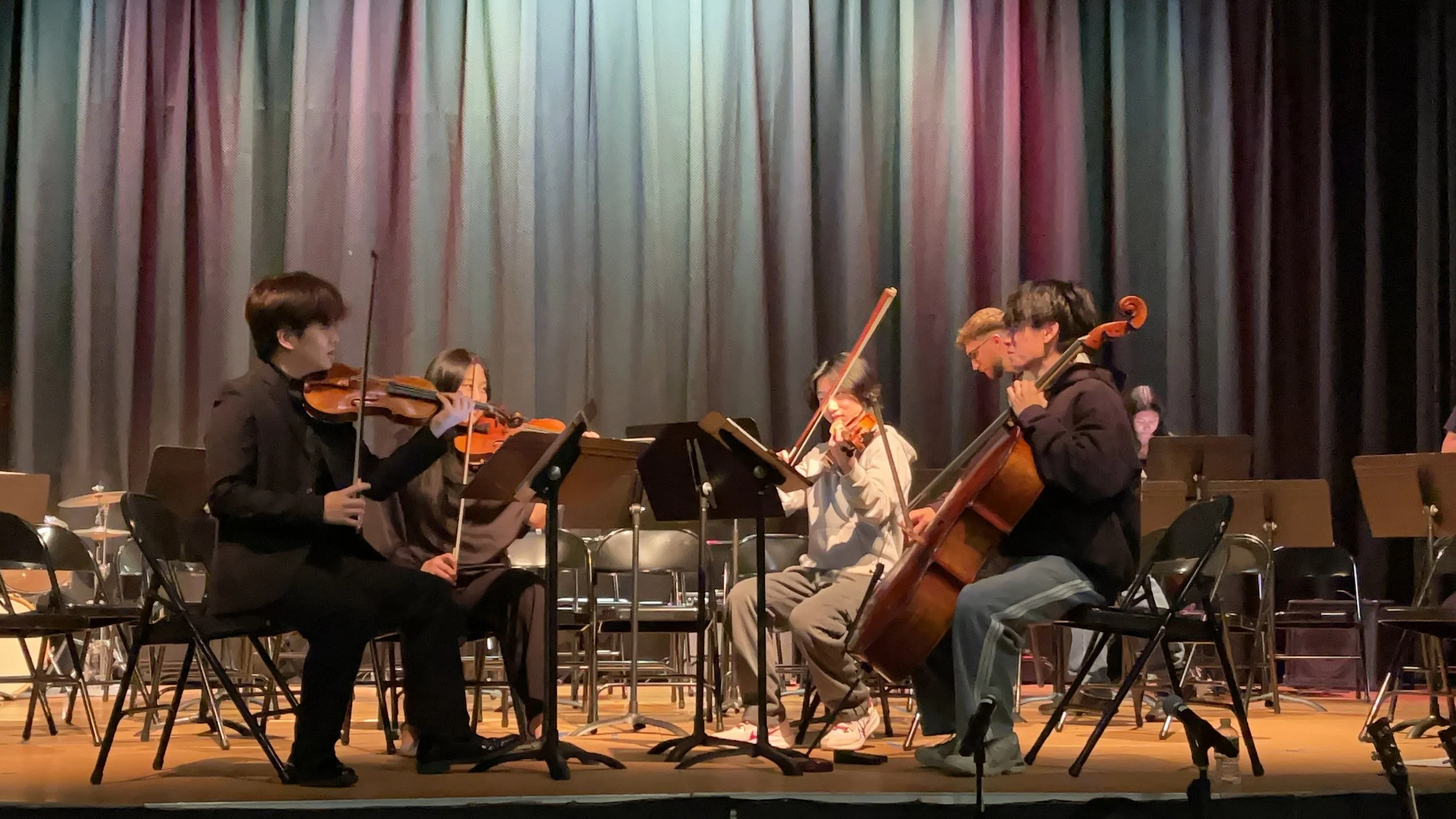 Young musicians playing string instruments, including violins and a cello, on a stage with a dark curtain backdrop.