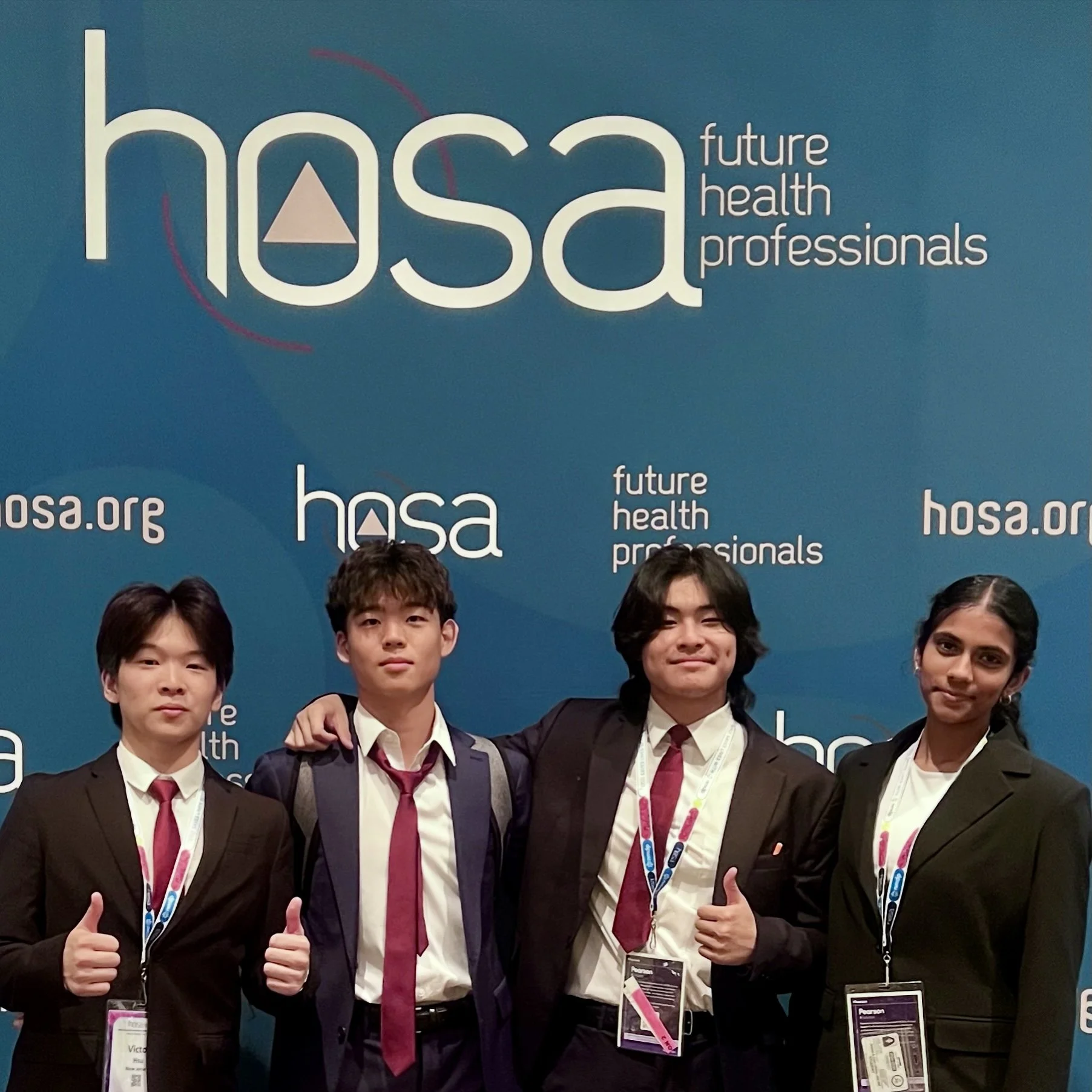 Four young professionals in formal attire at a conference, standing in front of a blue backdrop with the HOSA logo and tagline 'future health professionals', smiling and giving thumbs-up.