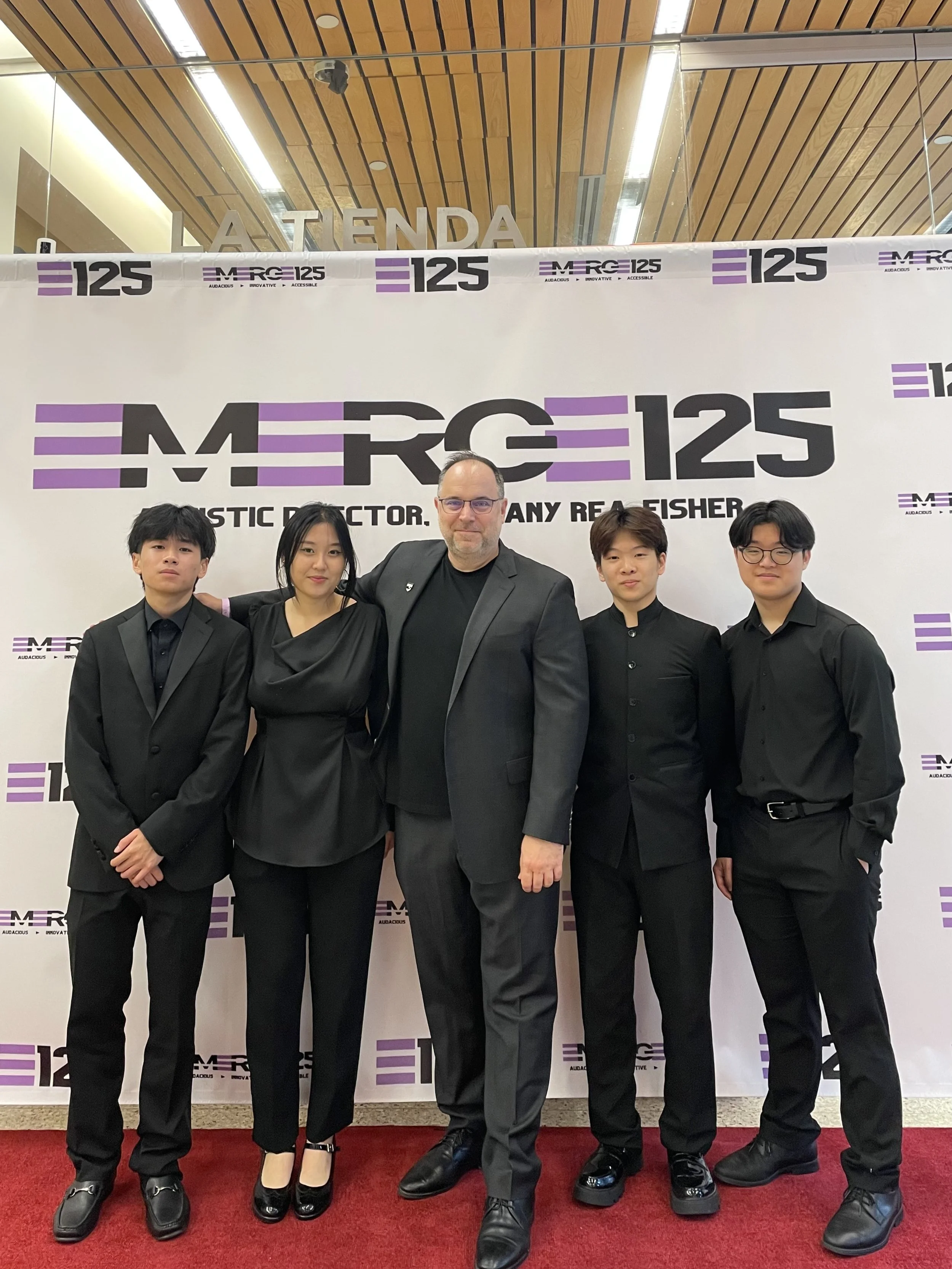 A group of five people, four men and one woman, dressed in black, standing together in front of a promotional backdrop with the text 'MERG 125' and 'Autistic Director. Any Real Finer'. They are at an event, standing on a red carpet inside a building 