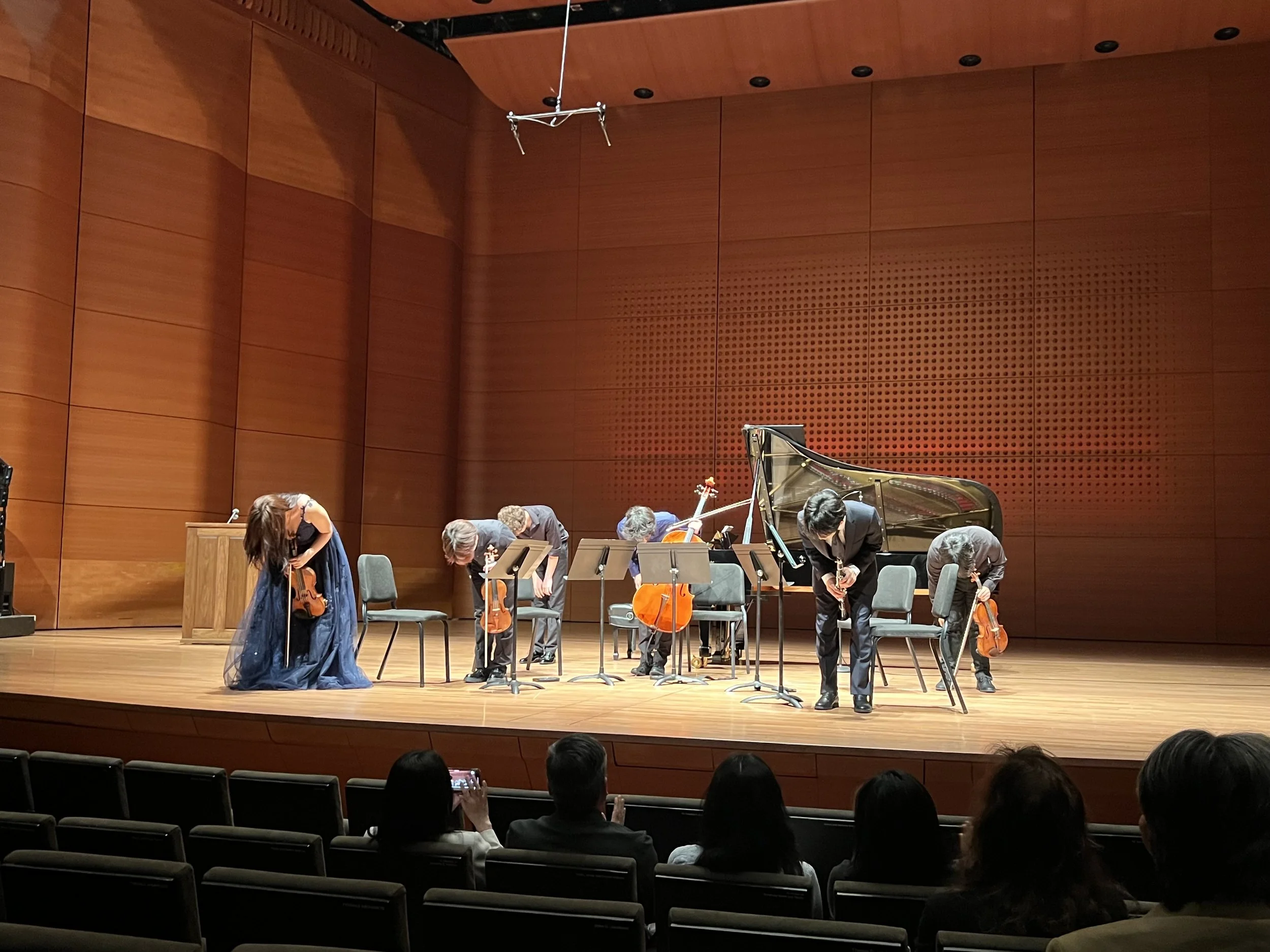 Orchestra on stage bowing after performance in concert hall with audience watching.