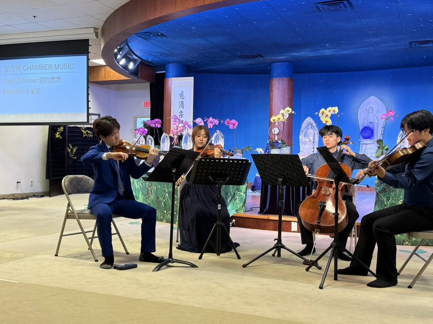 Four musicians playing string instruments on stage during a performance, with a backdrop of religious statues, orchids, and a large screen displaying information about the event.
