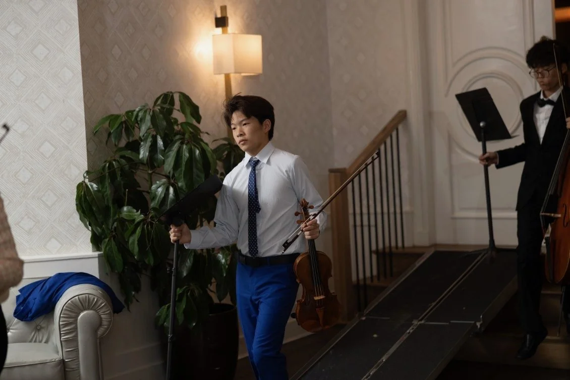 A young man in a white dress shirt, blue pants, and a navy polka-dot tie holding a violin and a bow, walking off a small stage in an elegant room with patterned wallpaper and a large green plant. Another person in formal attire is partially visible i