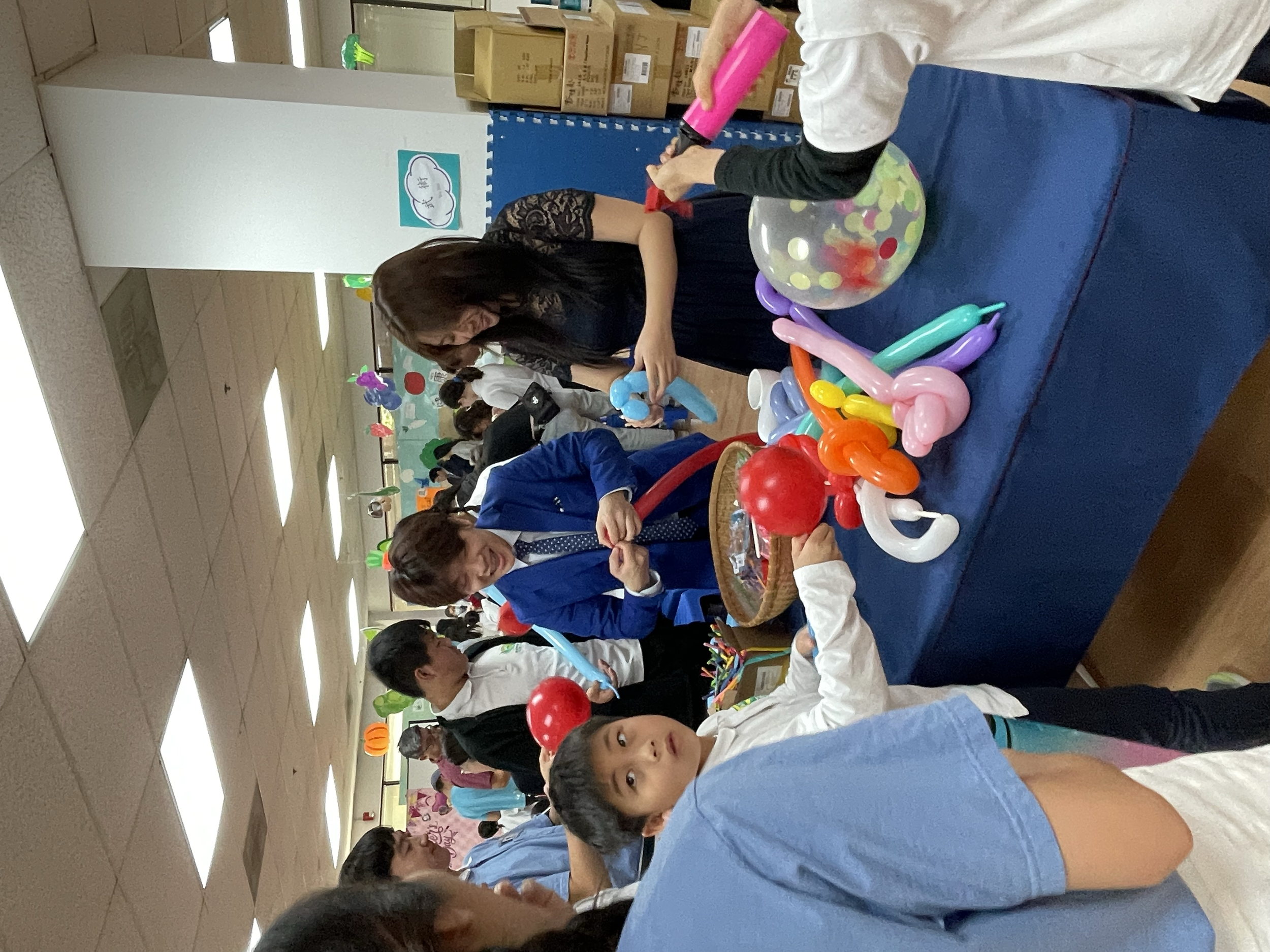 Children and adults at a balloon art station during a festive event, with balloons on a table and colorful decorations in the background.