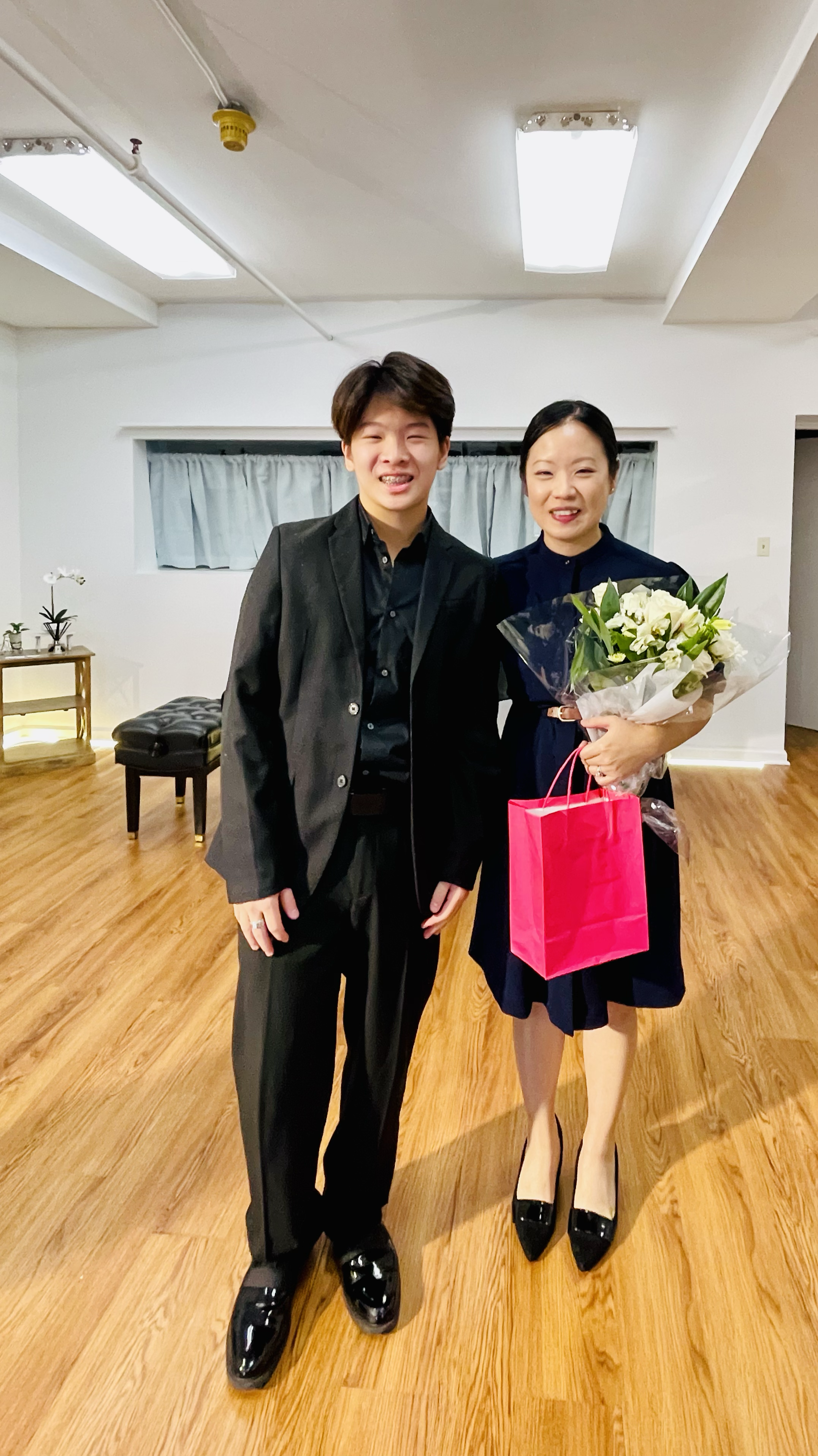 A young man and an older woman smiling and standing together in a room. The woman holds a bouquet of white flowers and a pink gift bag. The room has wooden floors, a white wall with a small window, a bench, and a table with a potted plant.