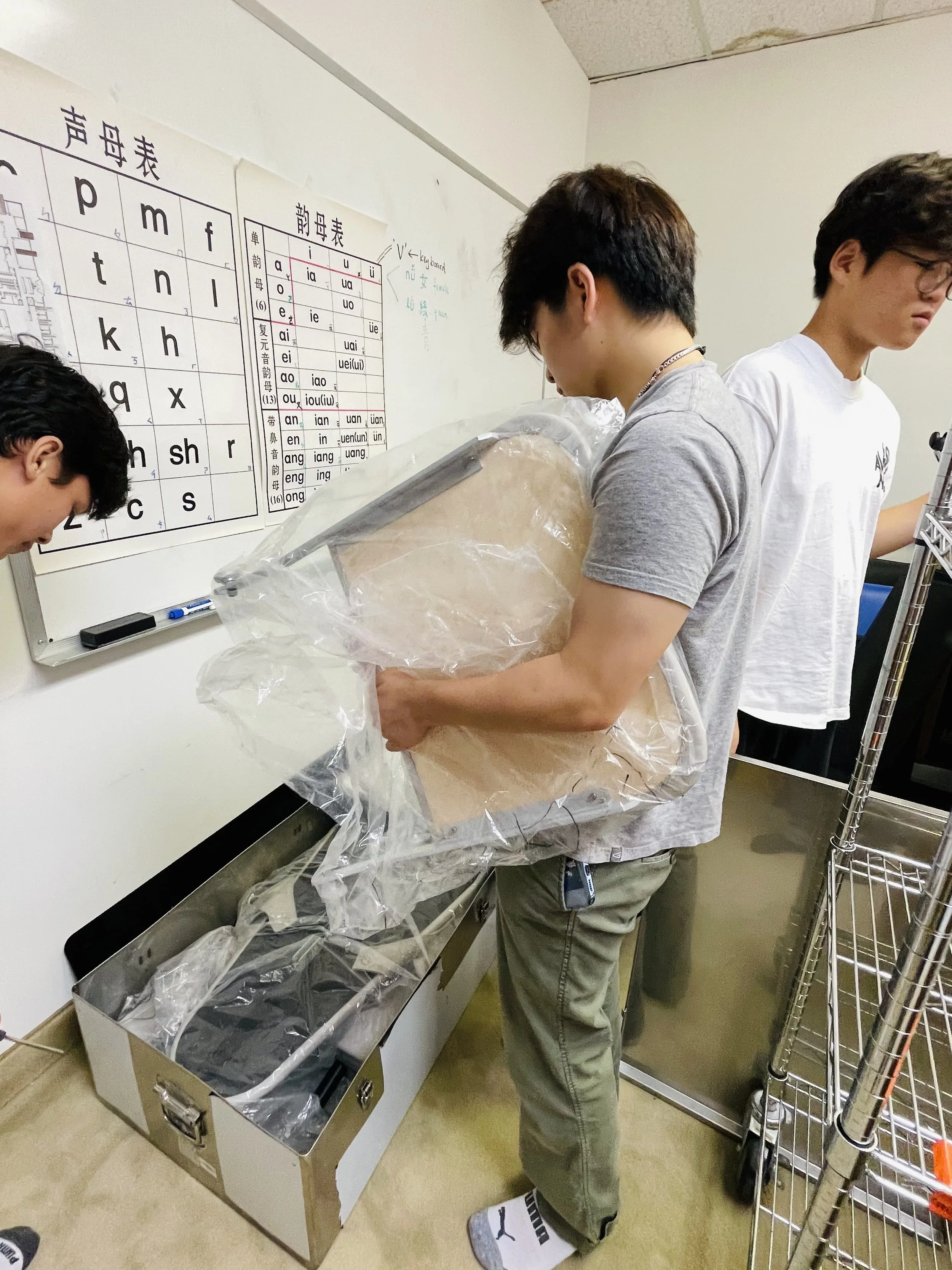 Three young men in a room with Japanese language charts on the wall. One man is holding a large object wrapped in plastic, standing near a metal box.
