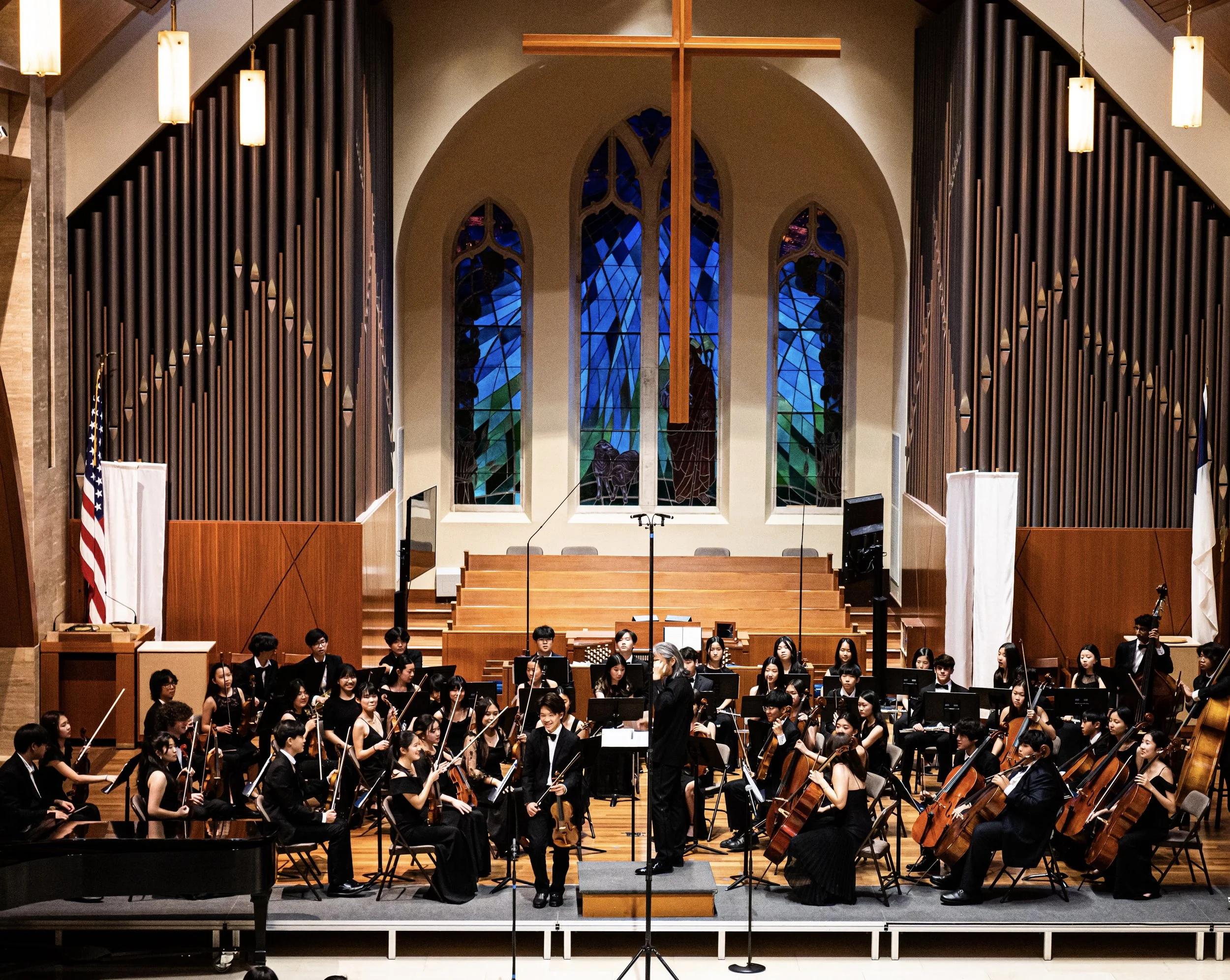 A symphony orchestra performing on stage inside a church with stained glass windows, wooden cross, and choir seating, led by a conductor.