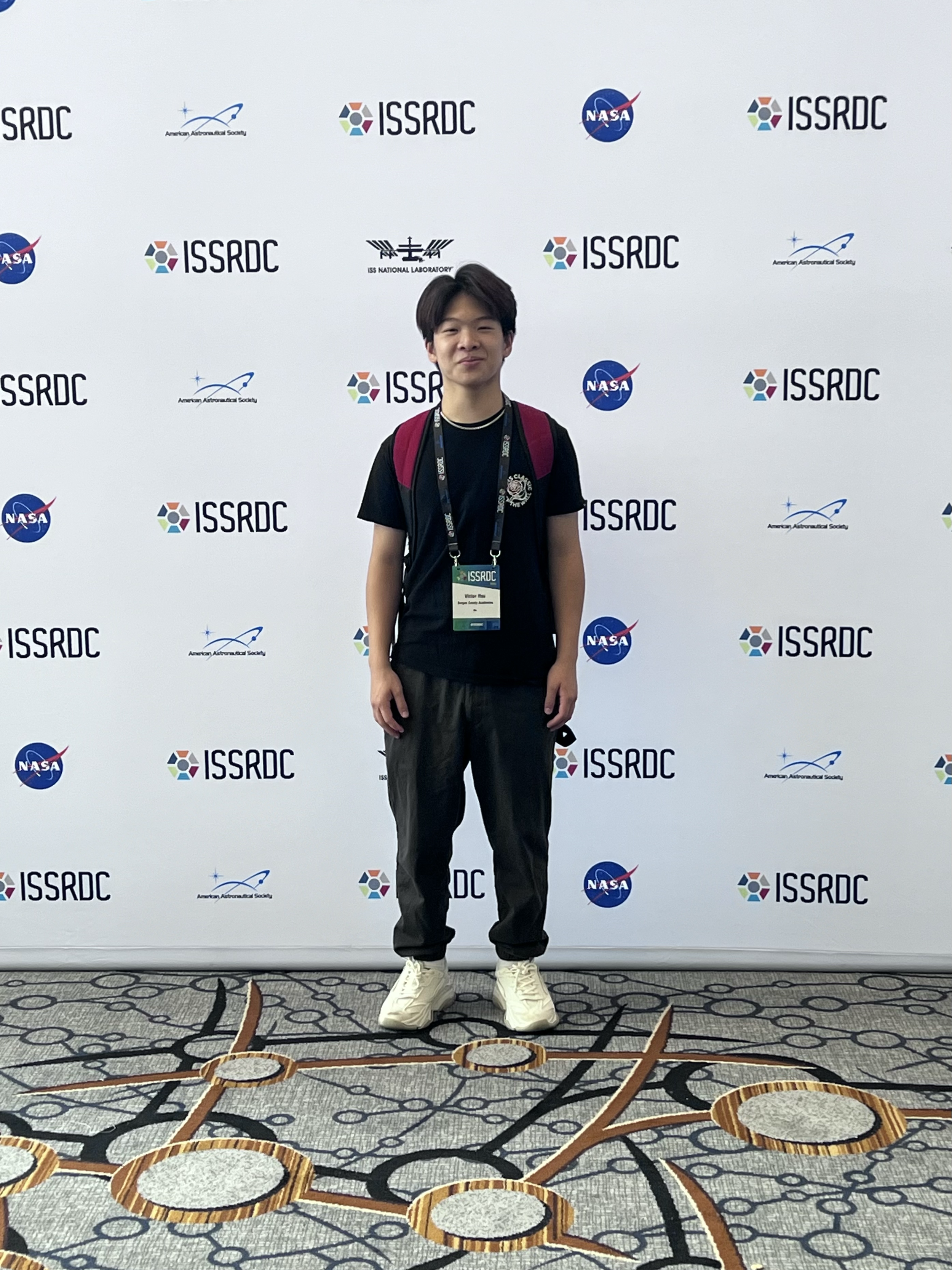 Young man standing in front of a backdrop with ISSRDC, NASA, and American Astronautical Society logos, wearing a black T-shirt, dark pants, white sneakers, and a lanyard with a name badge, on a patterned carpet.