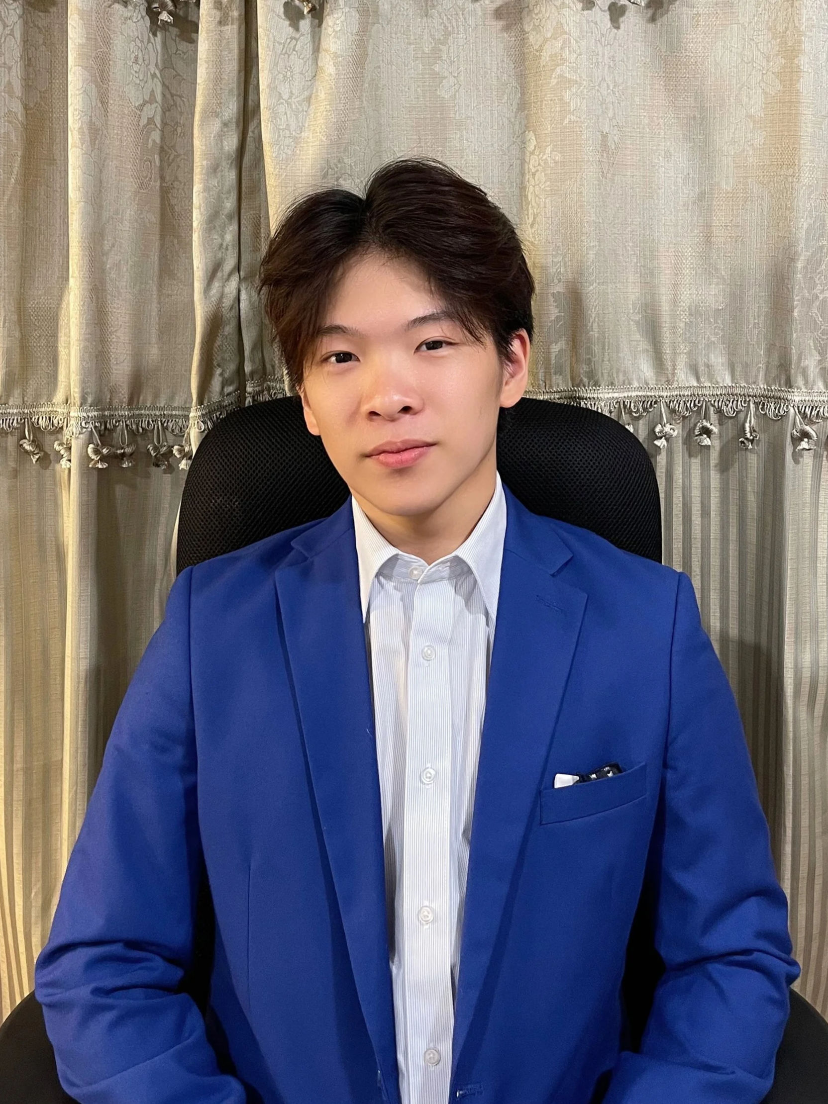 A young man with dark hair wearing a blue suit and white shirt, sitting in a black chair in front of beige curtains.
