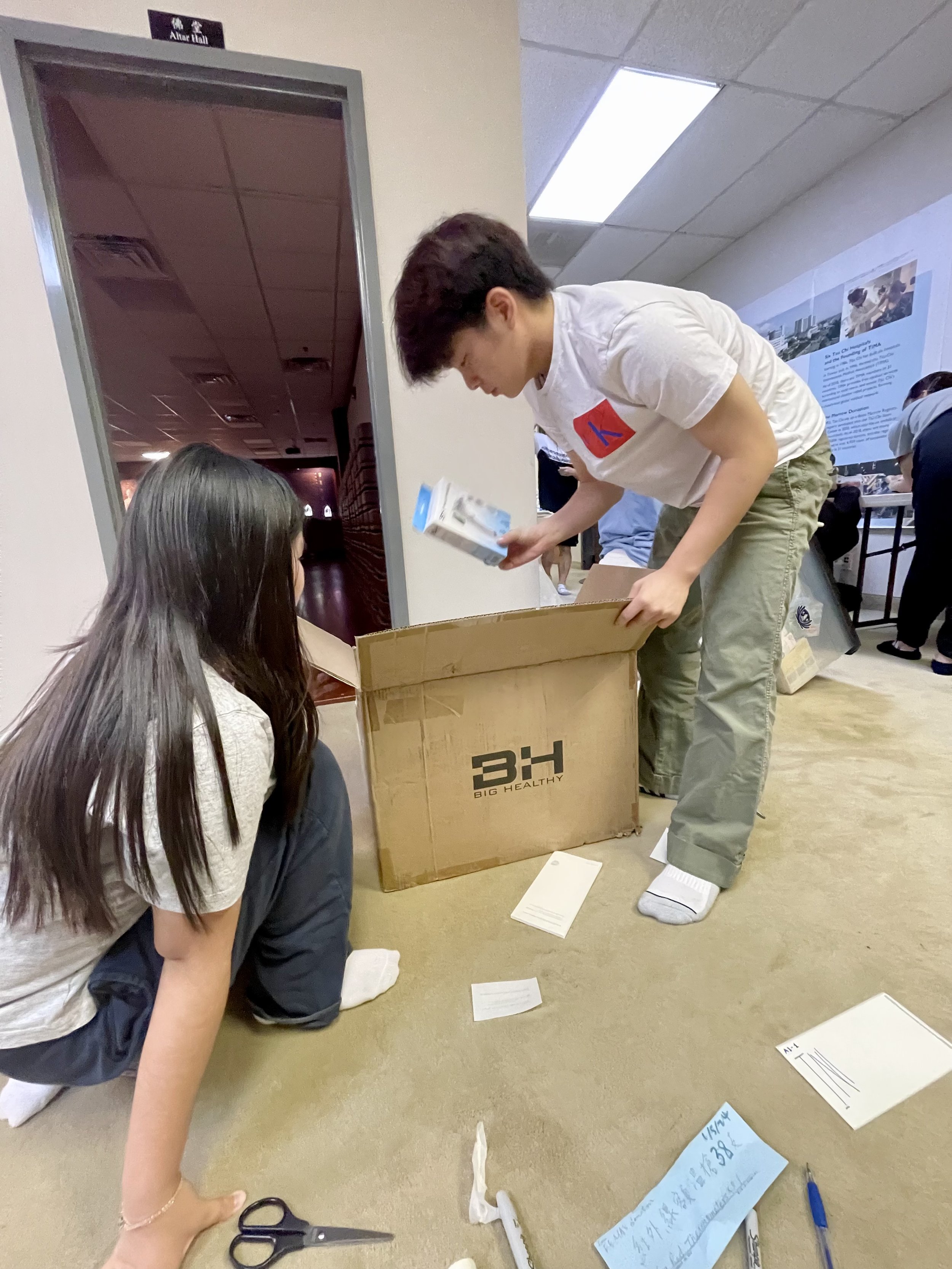 Two young individuals, a woman and a man, are engaged in unpacking or organizing items from a large cardboard box labeled 'BH BIG HEALTHY' on the floor of a room with carpeted flooring. The woman is kneeling on the floor, and the man is standing, hol