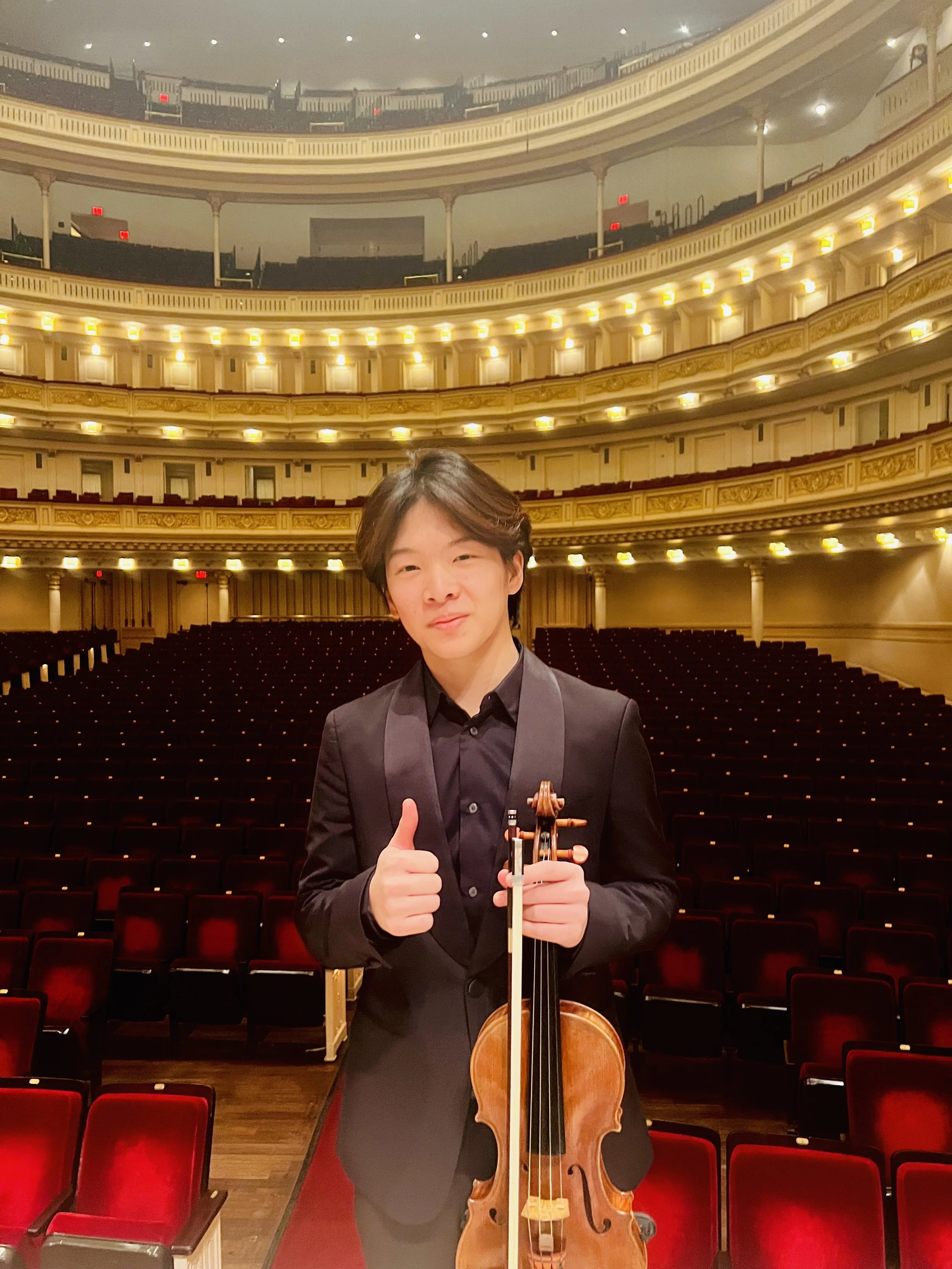A young man in a black suit standing on an empty concert hall stage, holding a violin and giving a thumbs-up gesture.