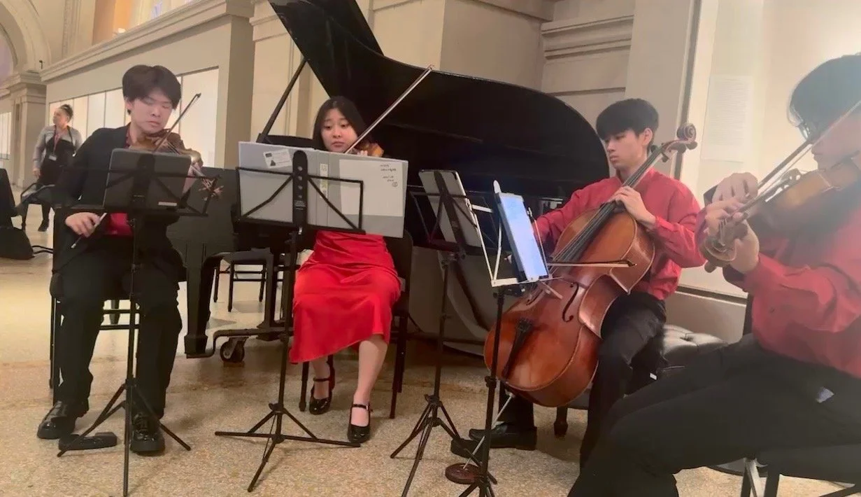 Four musicians playing string instruments, including two violinists, a cellist, and a pianist, in an indoor setting.