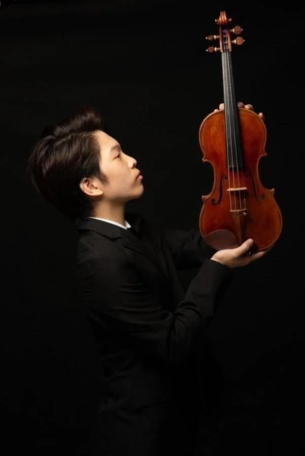 Young man in black suit holding a violin against a dark background