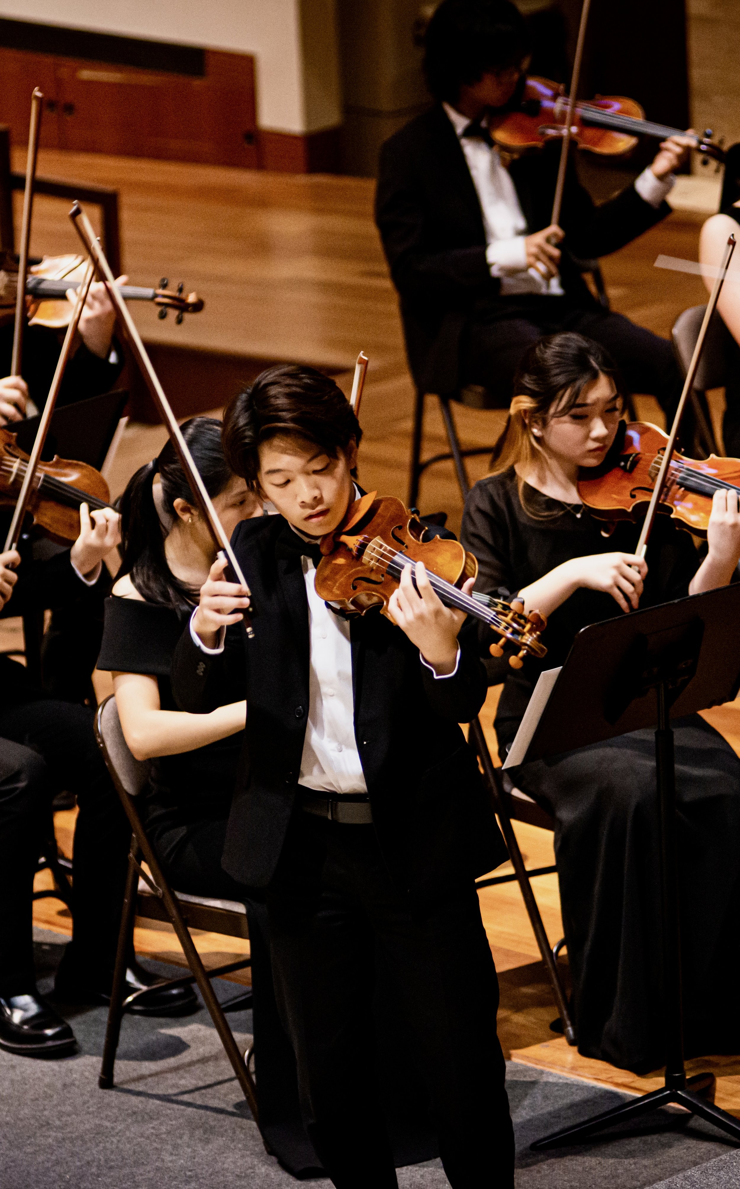 Young musicians in formal attire performing in an orchestra, playing violins on stage.