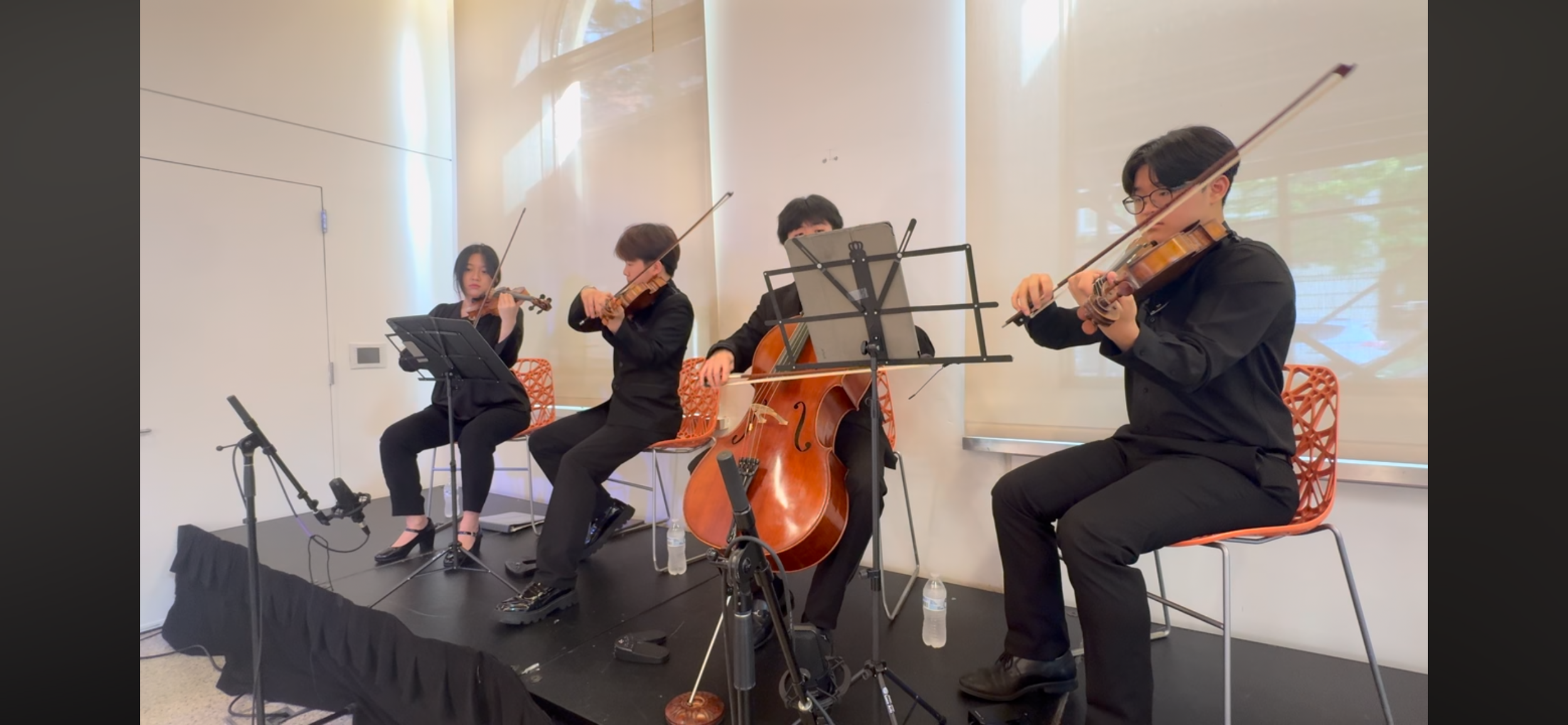Four musicians playing string instruments during a performance. From left to right: a woman with a violin, a man with a violin, a man with a cello, and another woman with a violin. They are seated on orange chairs and have music stands in front of th