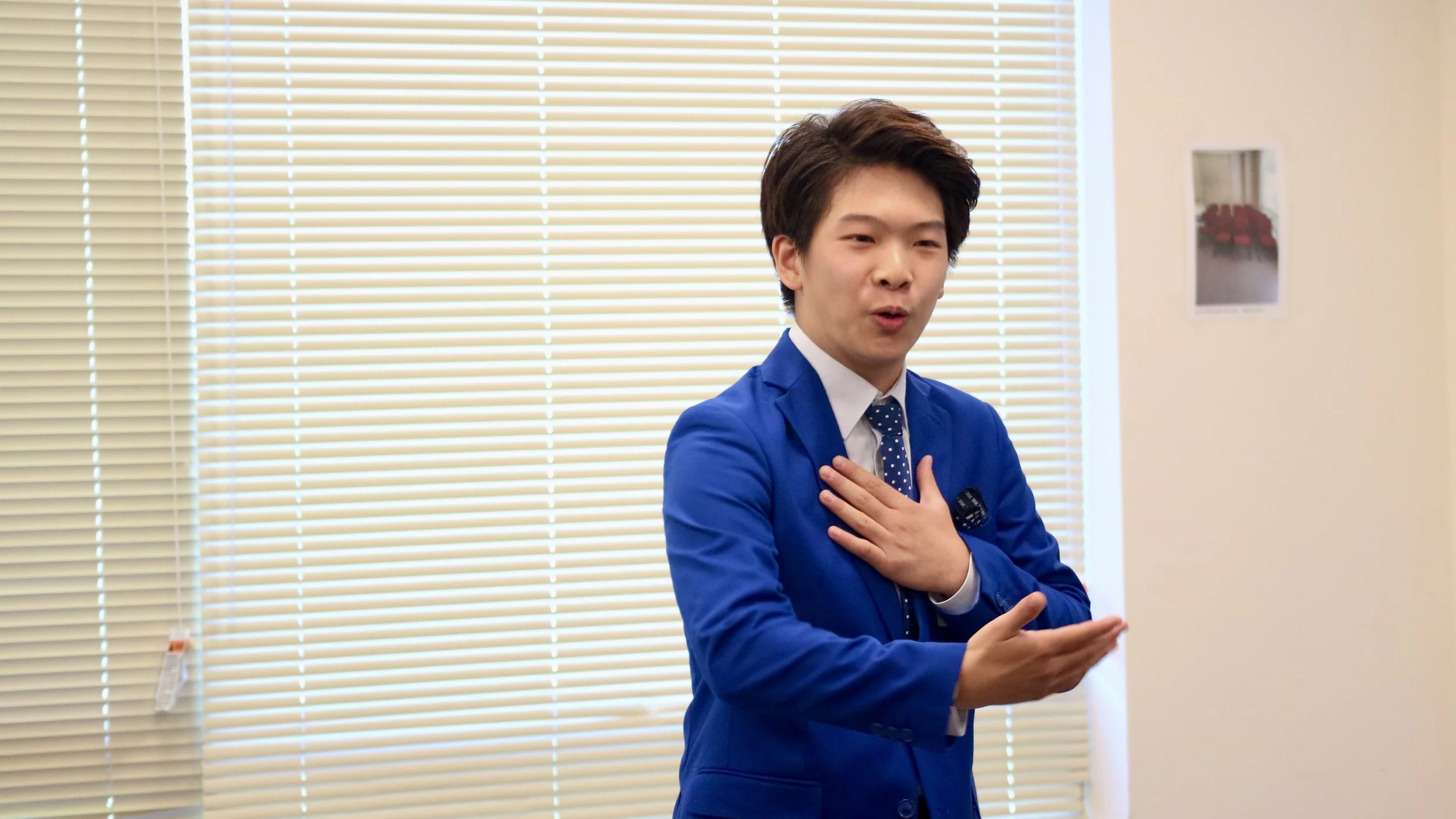 Young man in a blue suit with a polka dot tie, standing indoors in front of beige blinds, gesturing with his right hand and touching his chest with his left hand.