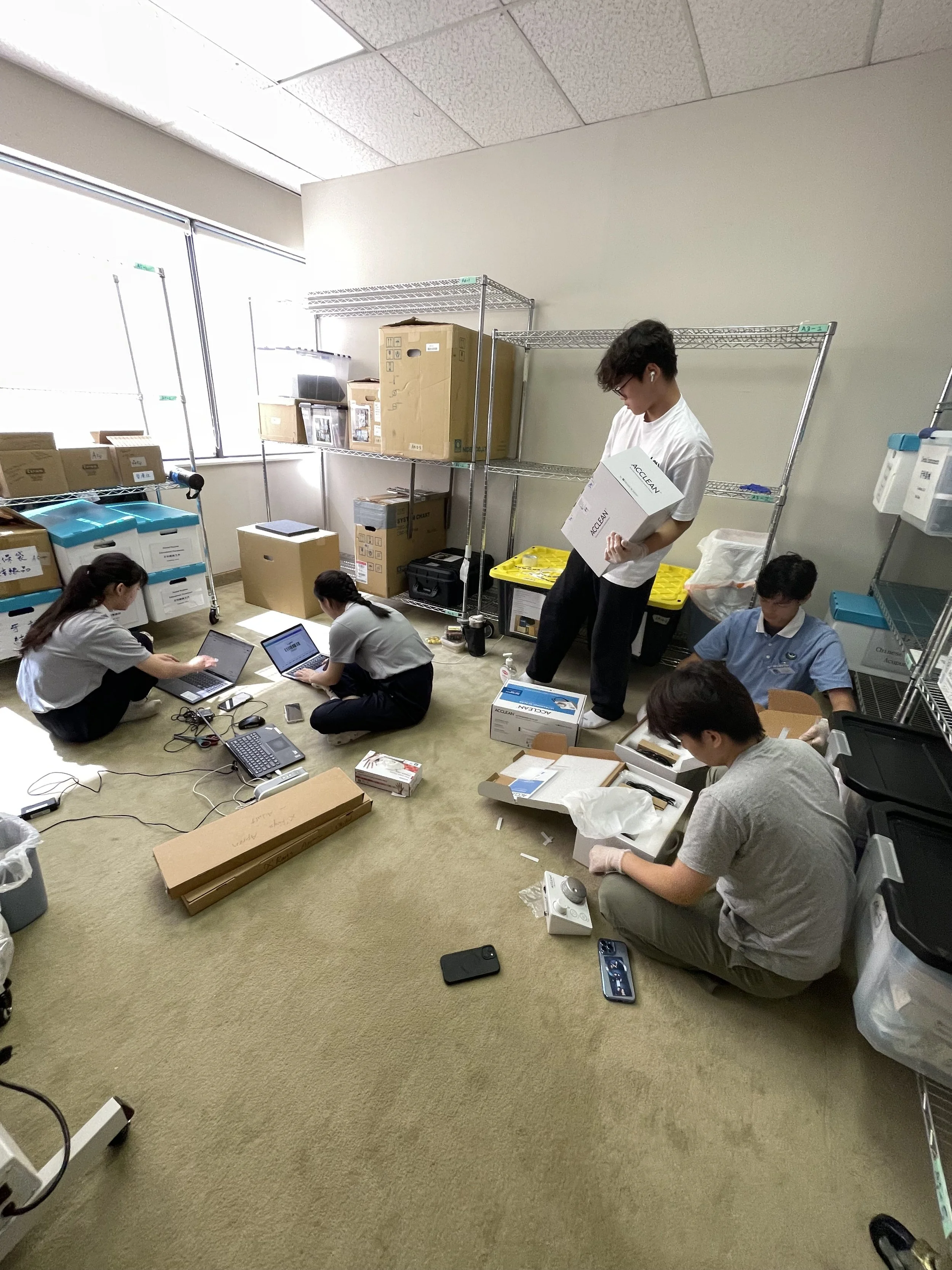 Group of five people working in a small office or storage room with laptops, boxes, and shelves.