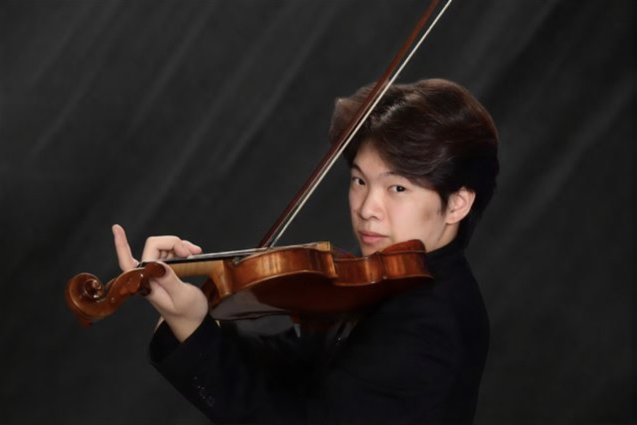 A young man with dark hair plays a violin, holding it with his left hand and using a bow with his right hand, against a dark background.