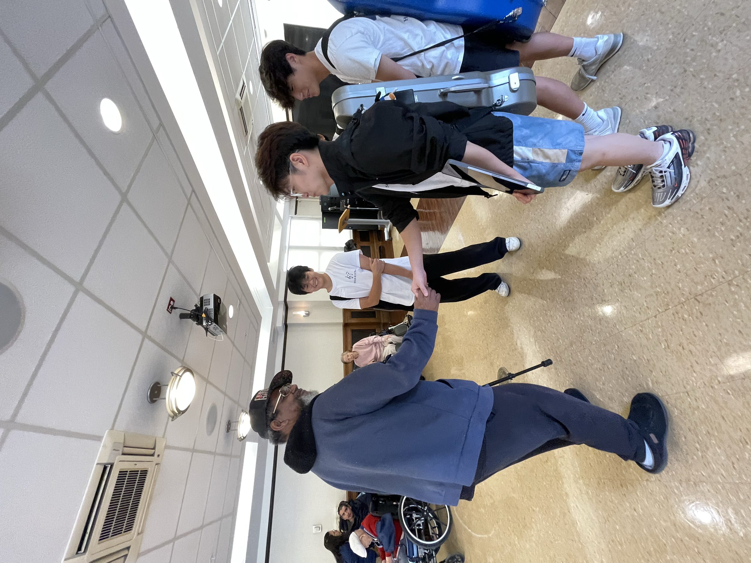 People standing in line while a man in a blue jacket and cap appears to be taking a photo or video of another person. Several people with luggage and backpacks are present in a room with a framed ceiling, bright lighting, and light-colored flooring.