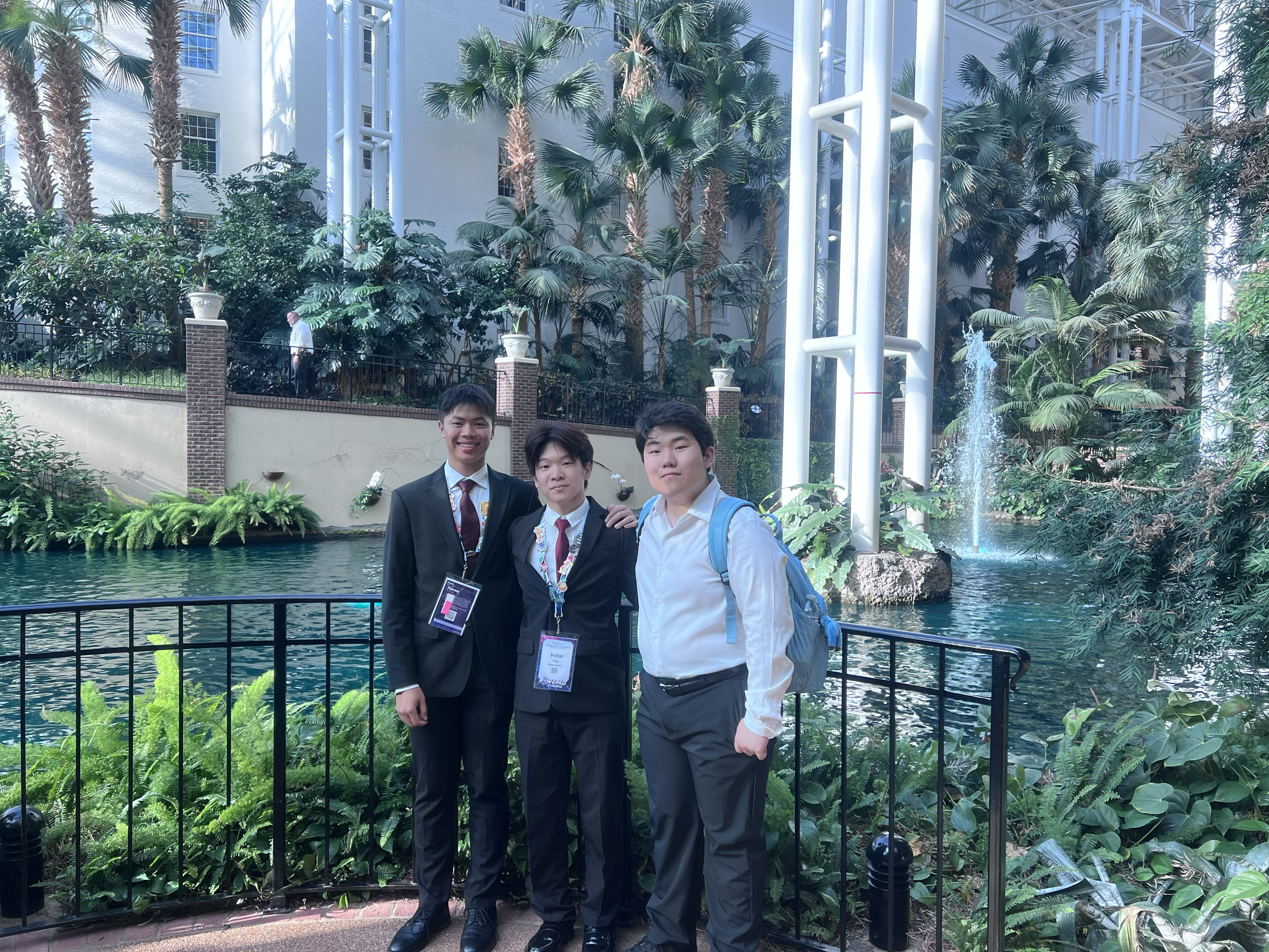 Three young men in suits standing in front of a water feature and lush greenery at an indoor venue, smiling and posing for the photo.