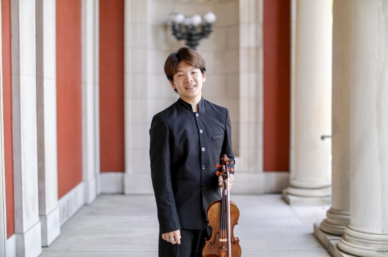 Young man in black standing in a columned corridor holding a violin and smiling.
