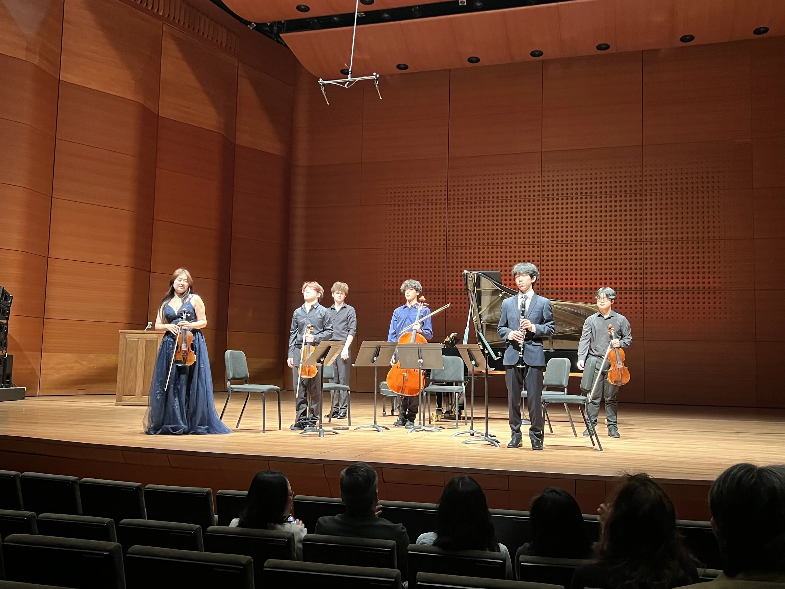 A group of six musicians on stage after a concert, with four holding violins, one holding a cello, and one standing near a piano, all dressed in formal attire, in a large concert hall with wooden walls and an audience in the foreground.