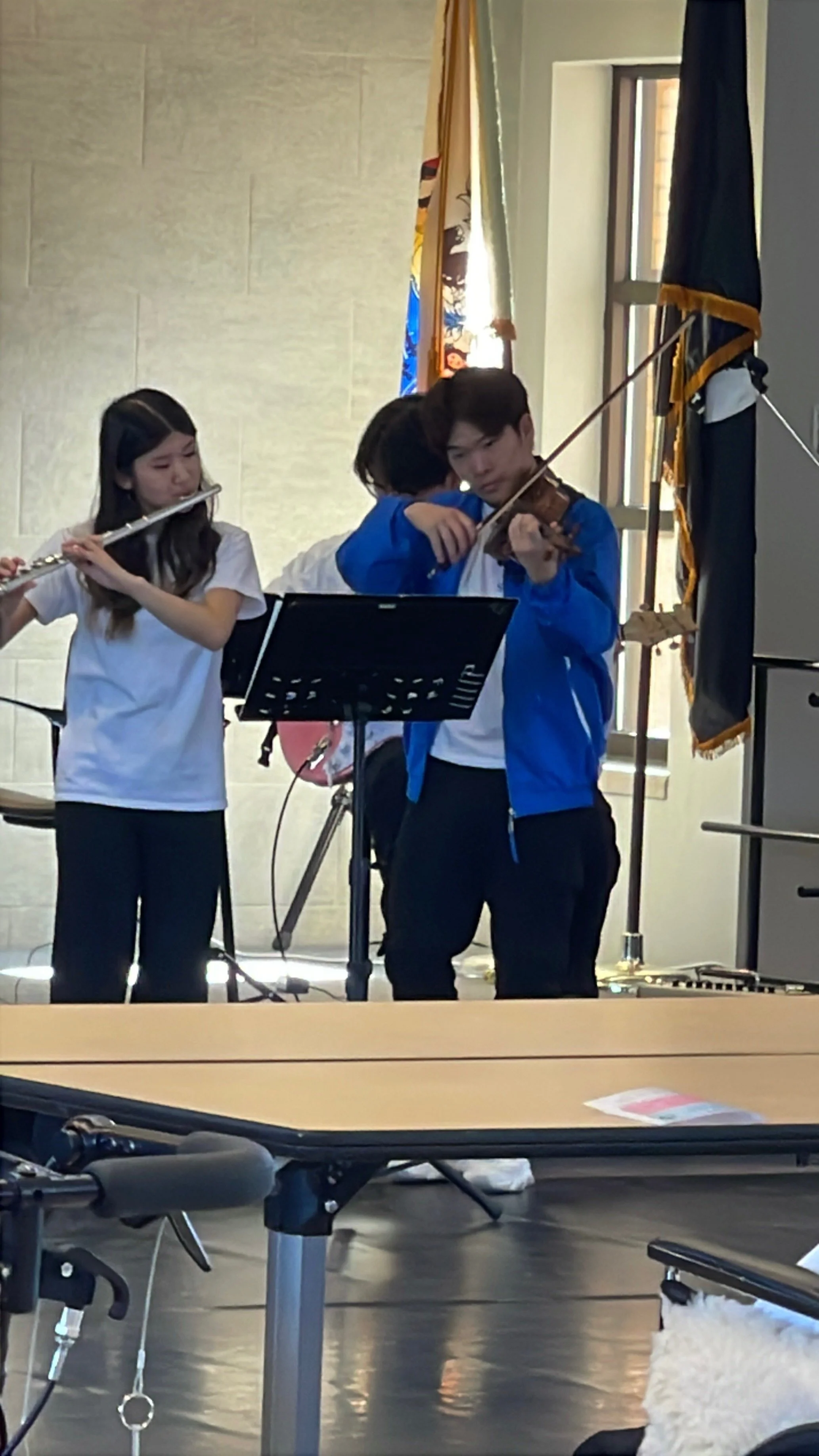 Young students playing musical instruments, including a violin and flute, during a performance or practice in a school or community room.