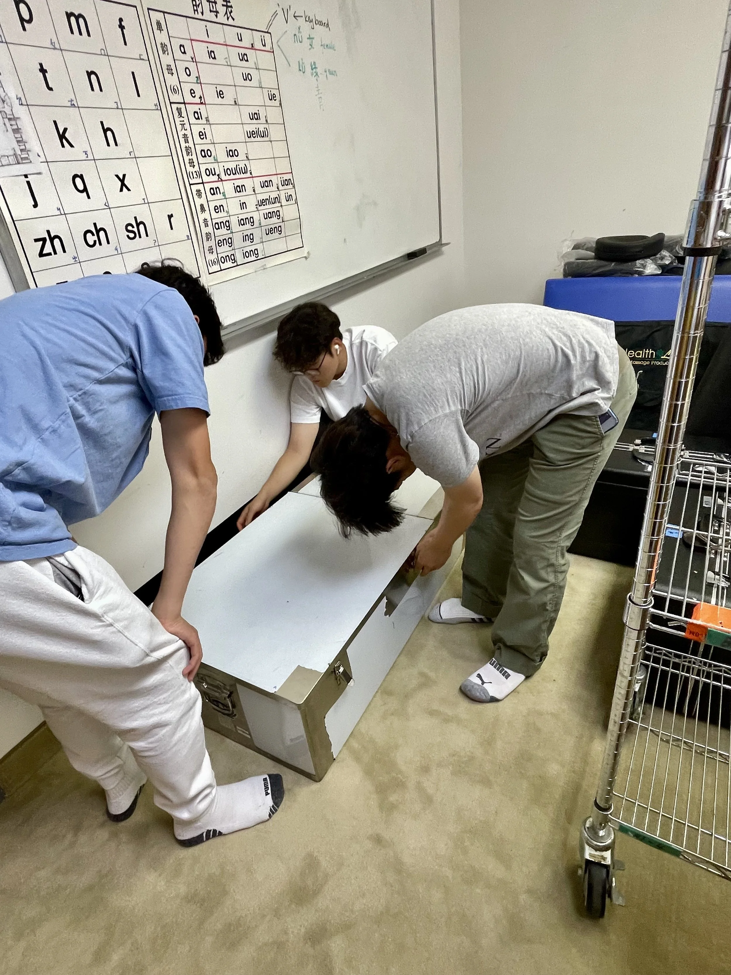 Three people working together on a project or repair in a room, with a whiteboard and a chart on the wall, a shopping cart on the right, and a white cabinet on the floor.