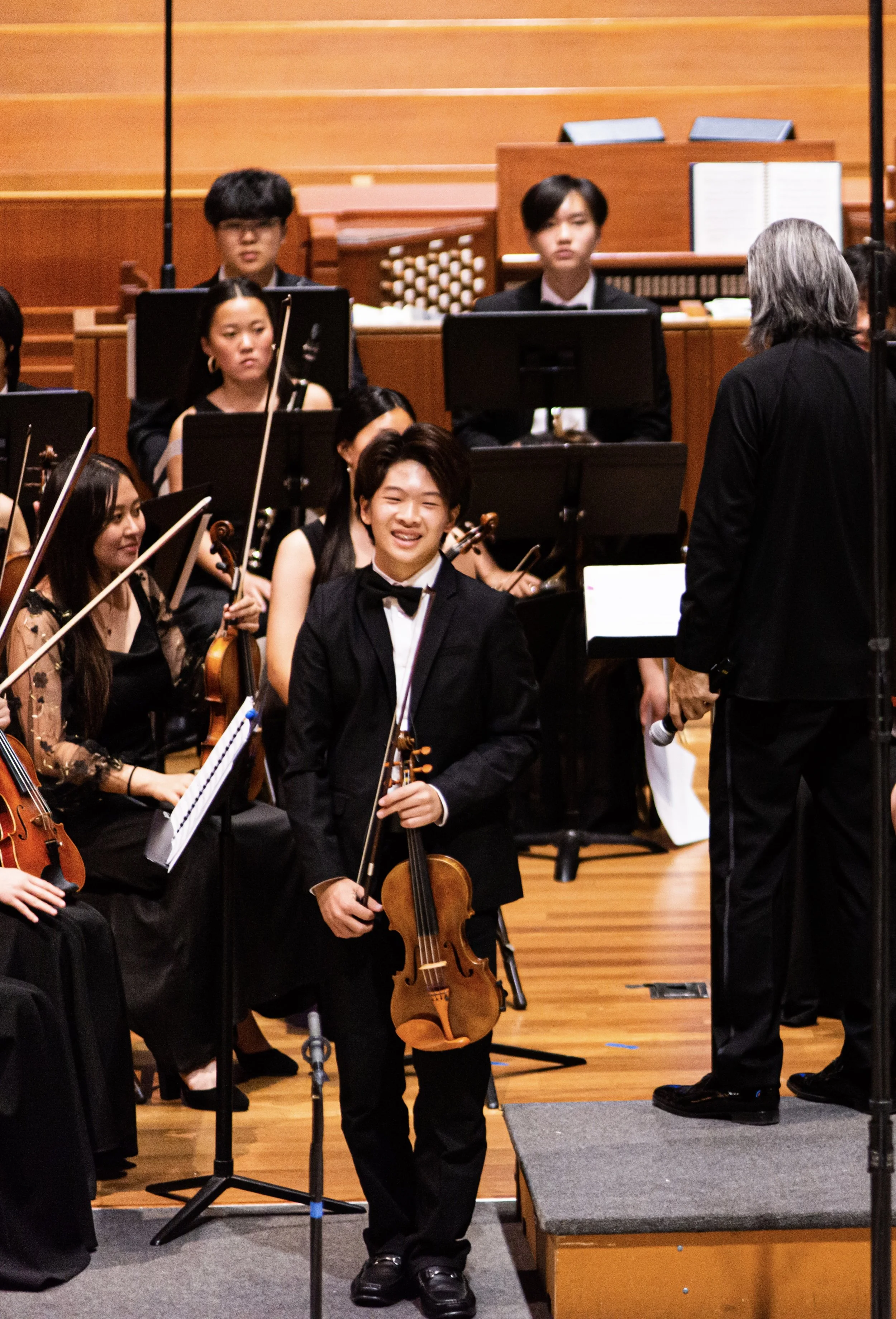 Young boy in tuxedo holding a violin, smiling on stage after an orchestra performance, with musicians and conductor in the background.