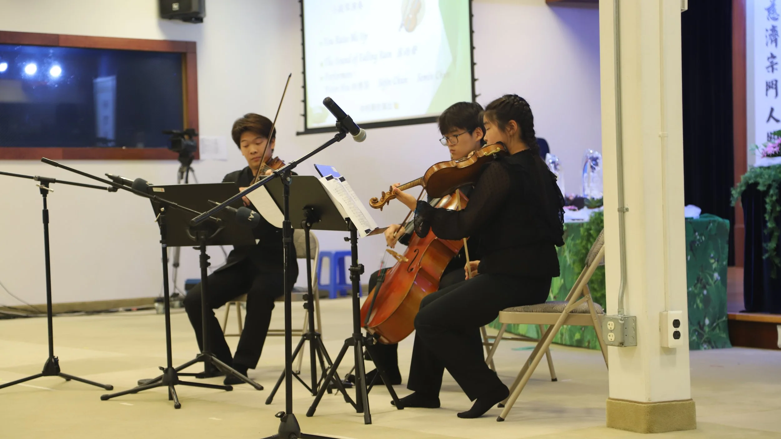 Three musicians playing string instruments: two on violins and one on cello, performing in an indoor setting with microphones and music stands.