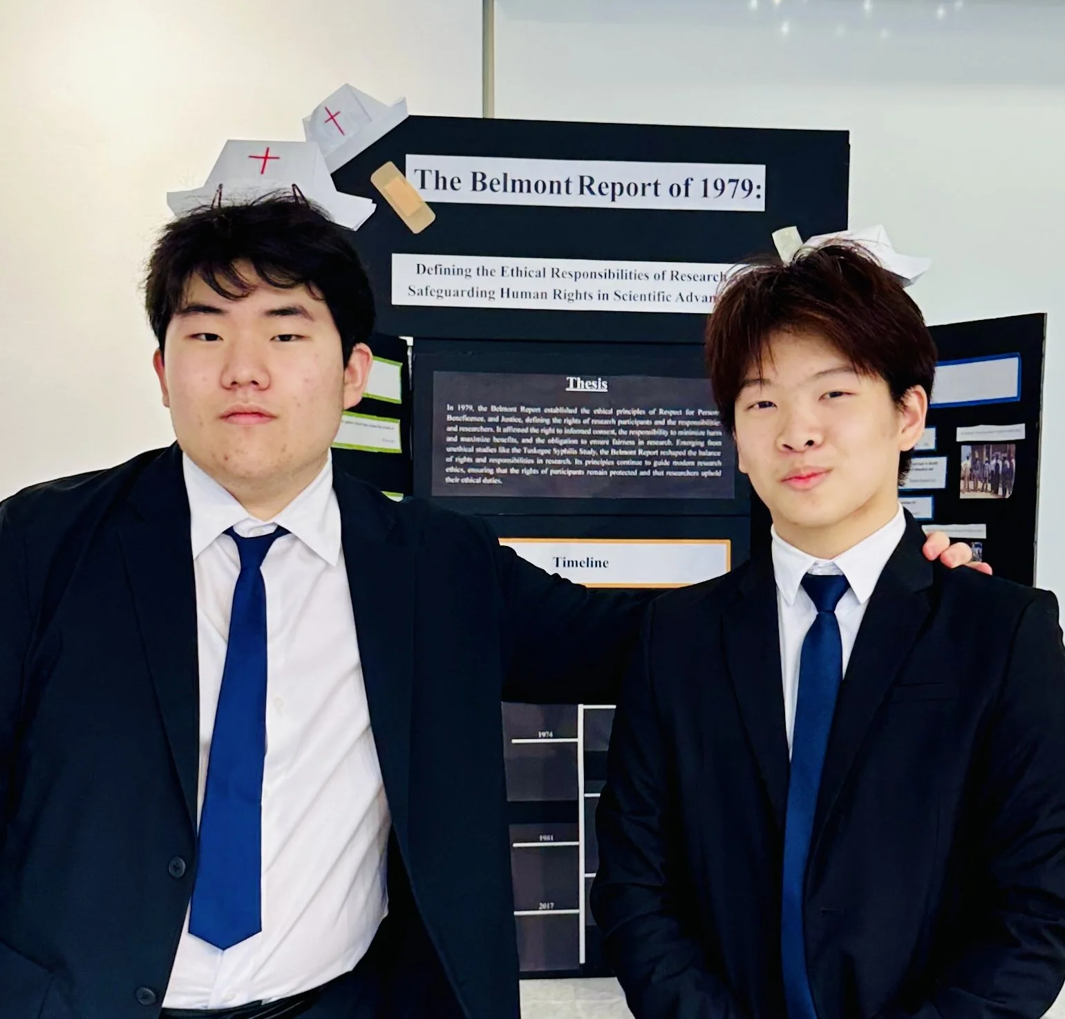 Two young men in formal suits with blue ties standing in front of a science research poster at a school or conference.
