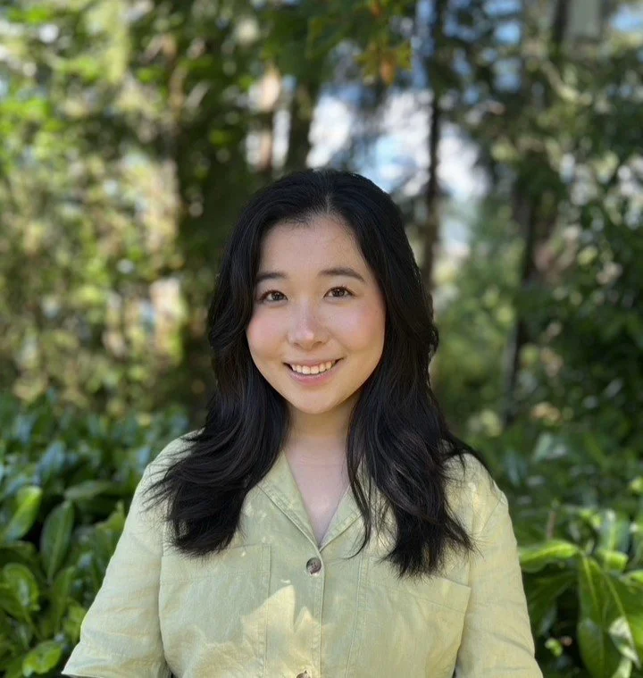 A young woman with long black hair smiling outdoors, wearing a light yellow shirt, with trees and greenery in the background.