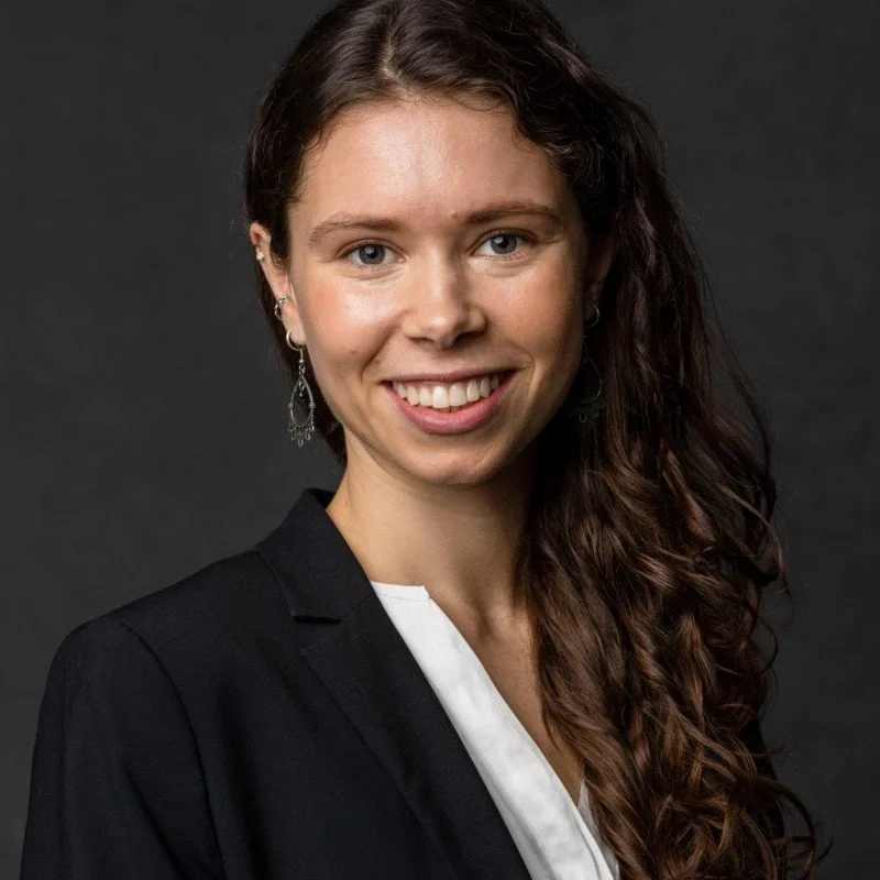 A young woman with long curly brown hair, fair skin, and blue eyes, smiling and wearing a black blazer with a white shirt, against a dark background.