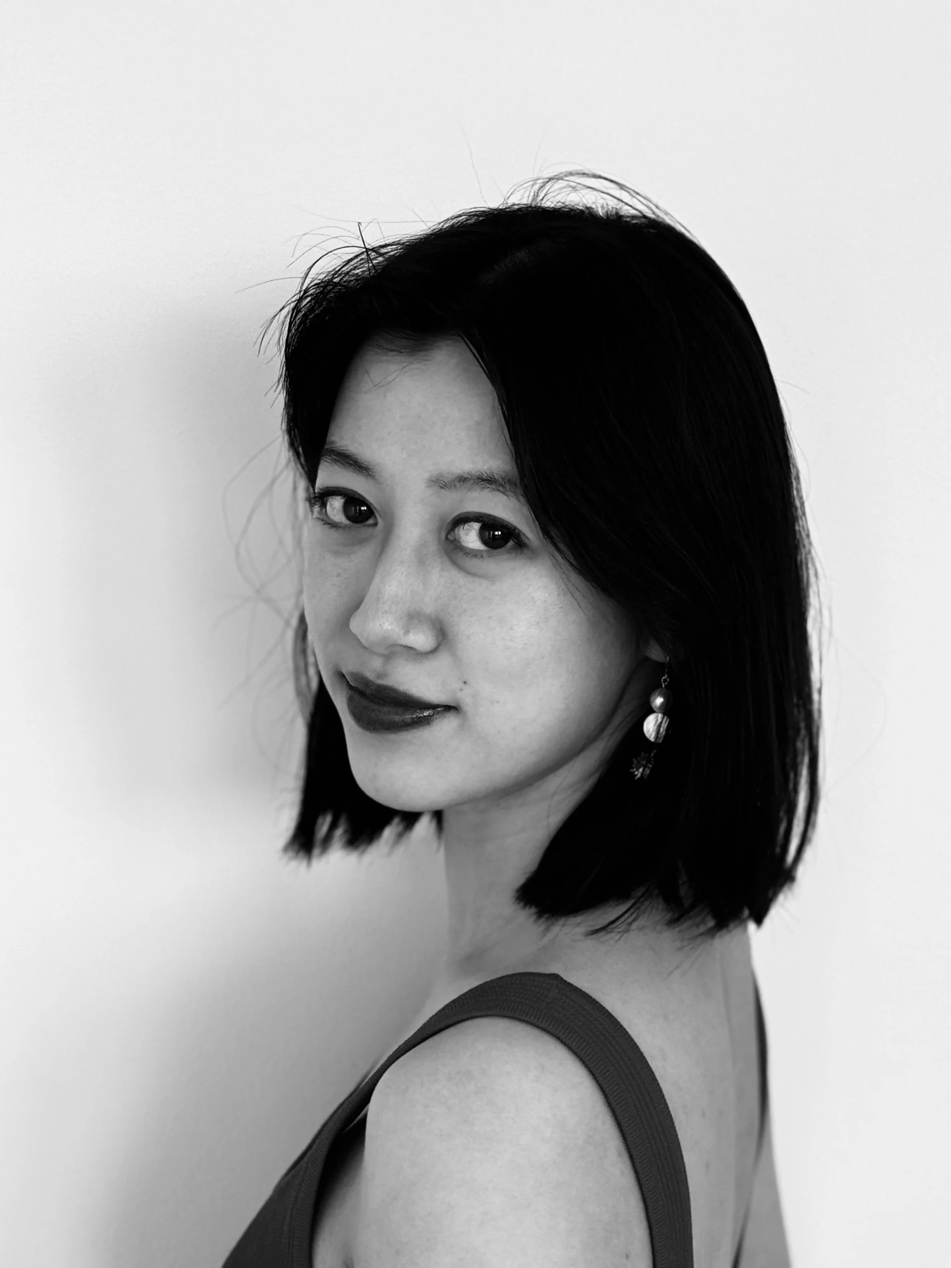 A black and white portrait of a woman with shoulder-length dark hair, wearing earrings and a sleeveless top, facing slightly to the left and looking at the camera.