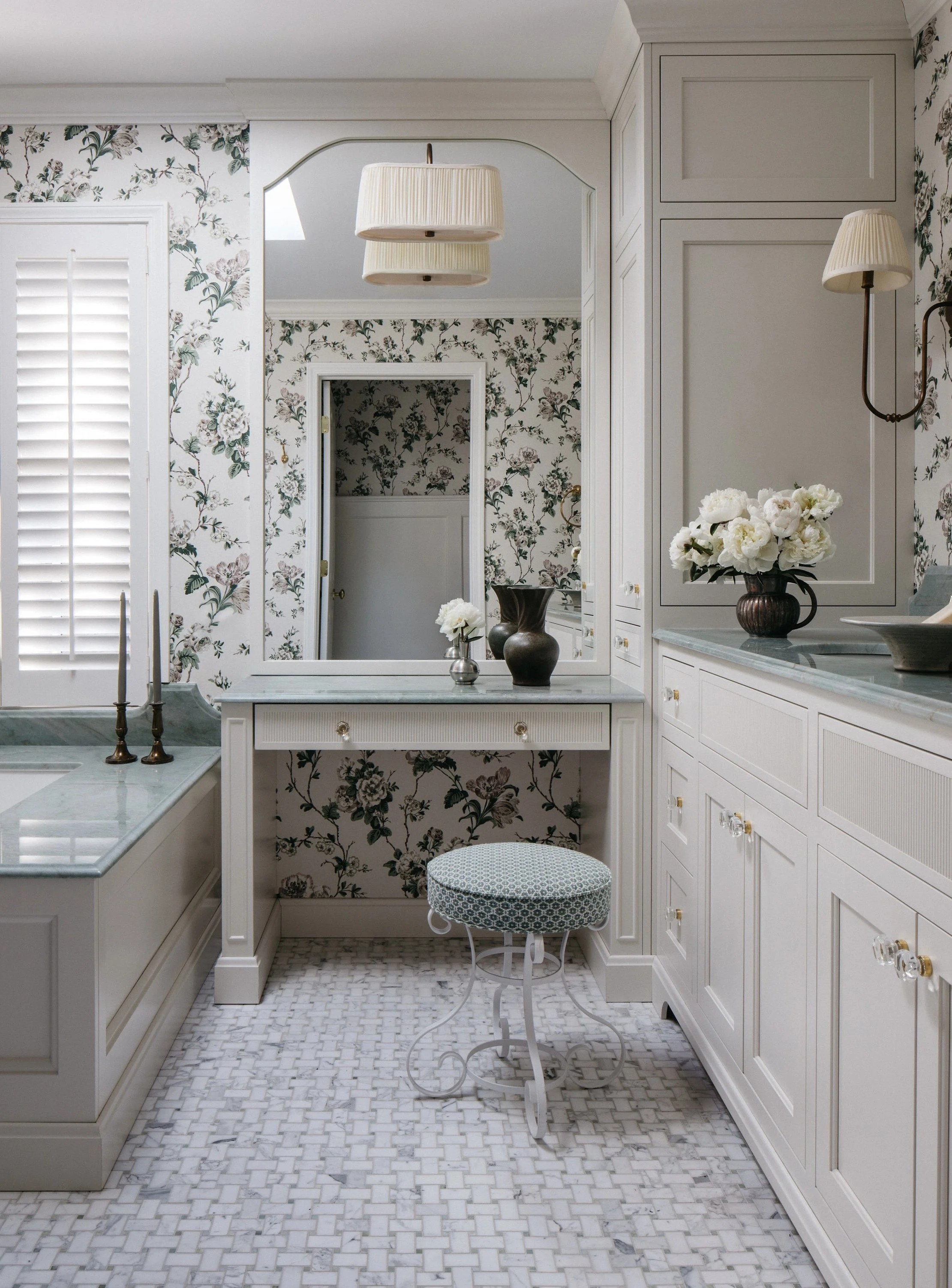 A vintage-style bathroom with floral wallpaper, white cabinetry, a marble countertop, a large mirror, a floral arrangement, and a small stool with a patterned cushion.