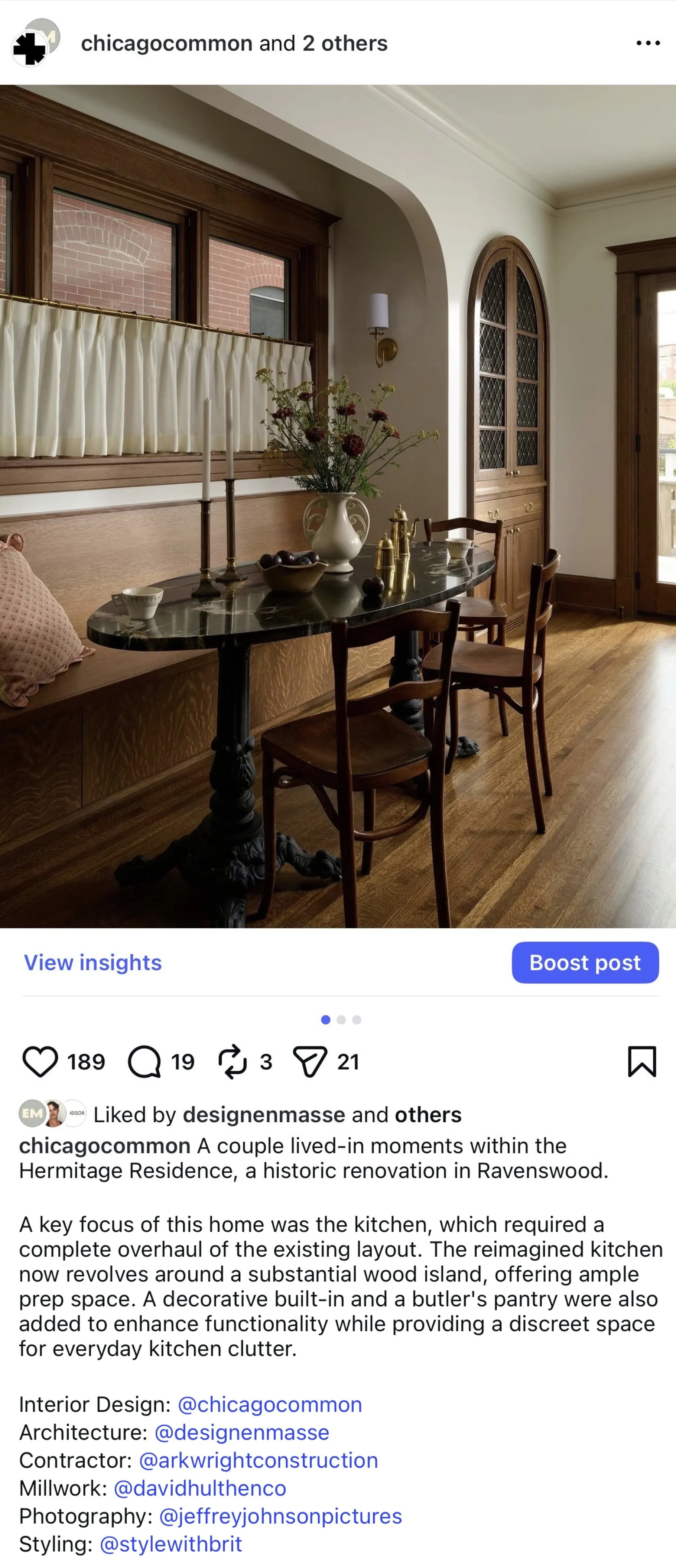 Interior showing a dining area with a wooden built-in bench, a black marble table with carved legs, three wooden chairs, a window with cream curtains, a decorative cabinet with glass doors, a door leading outside, a vase with flowers, candleholders, and small decorative items.