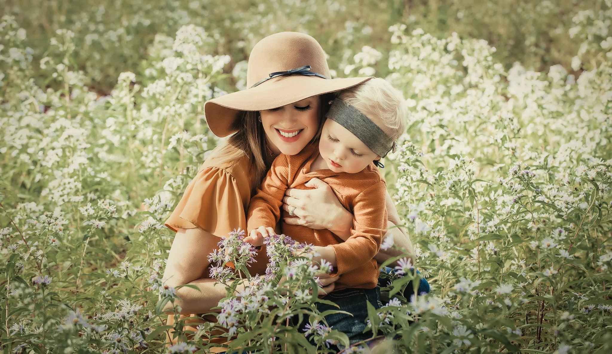 A smiling woman with a wide-brimmed hat holding a young boy in a field of purple and white flowers.