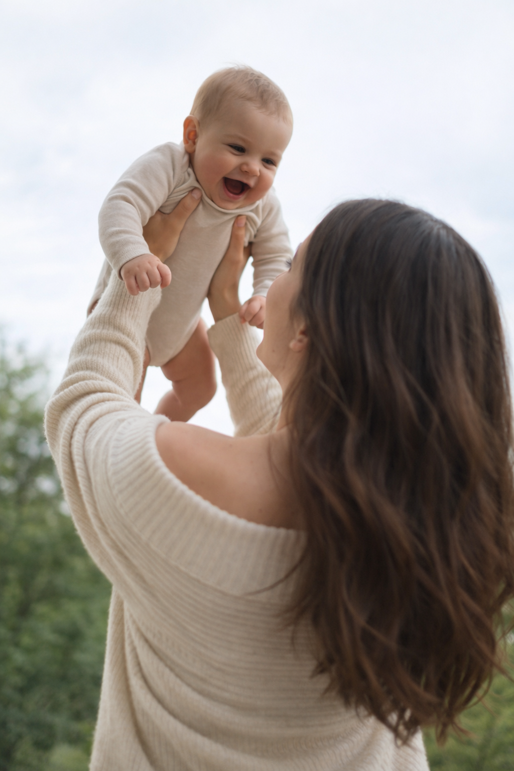 Woman lifting a smiling baby outdoors on a cloudy day.